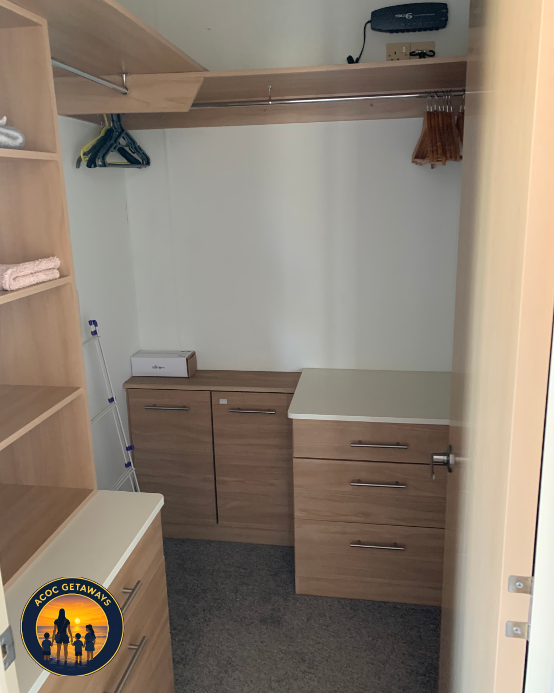 Empty wooden closet with hanging rod, drawers, and a small shelf, containing a few hangers, a box, and a yardstick, with a pair of shoes on an upper shelf and an electric socket on the wall.