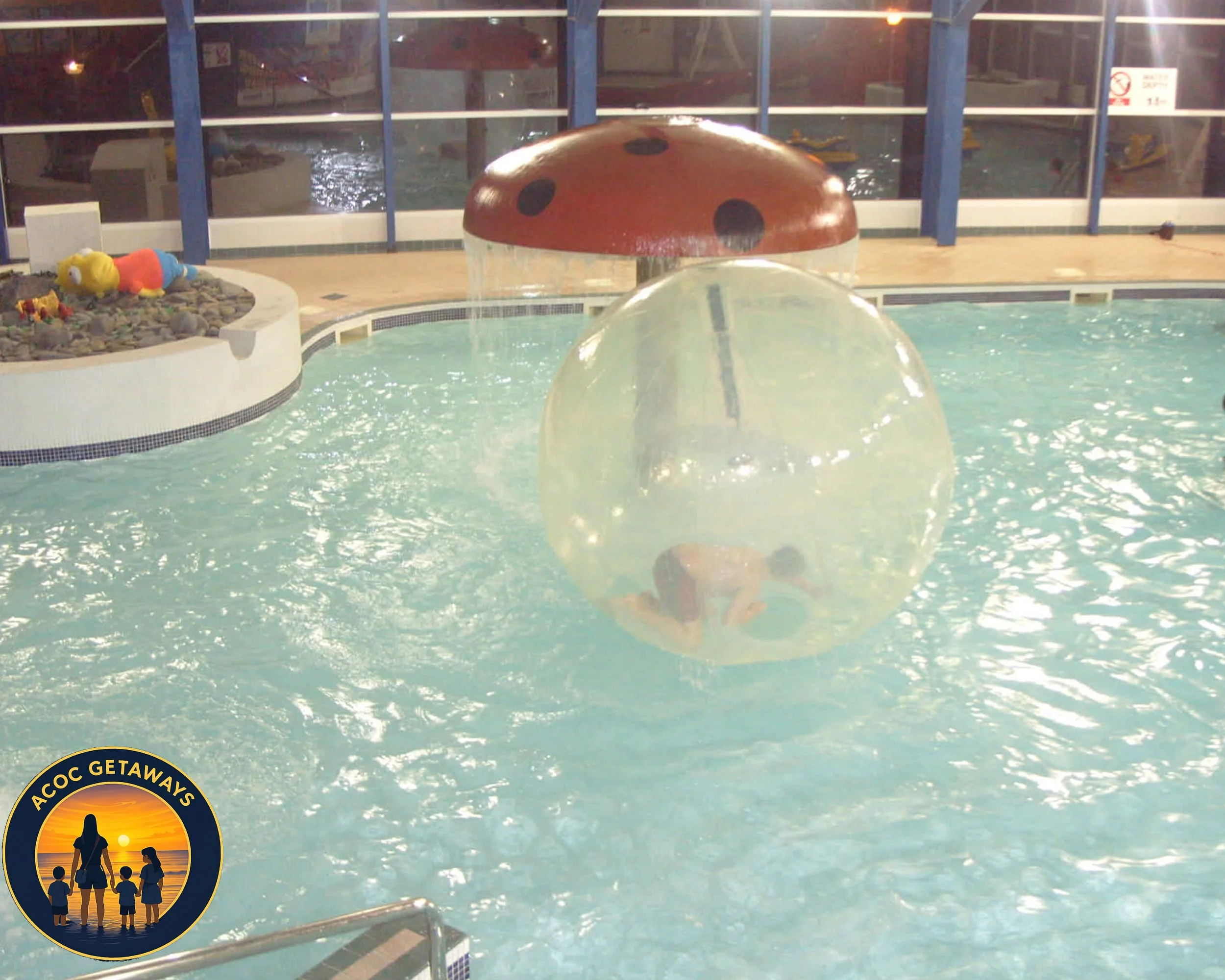 Person inside a large transparent inflatable ball floating on a swimming pool with a mushroom-shaped water fountain above it.