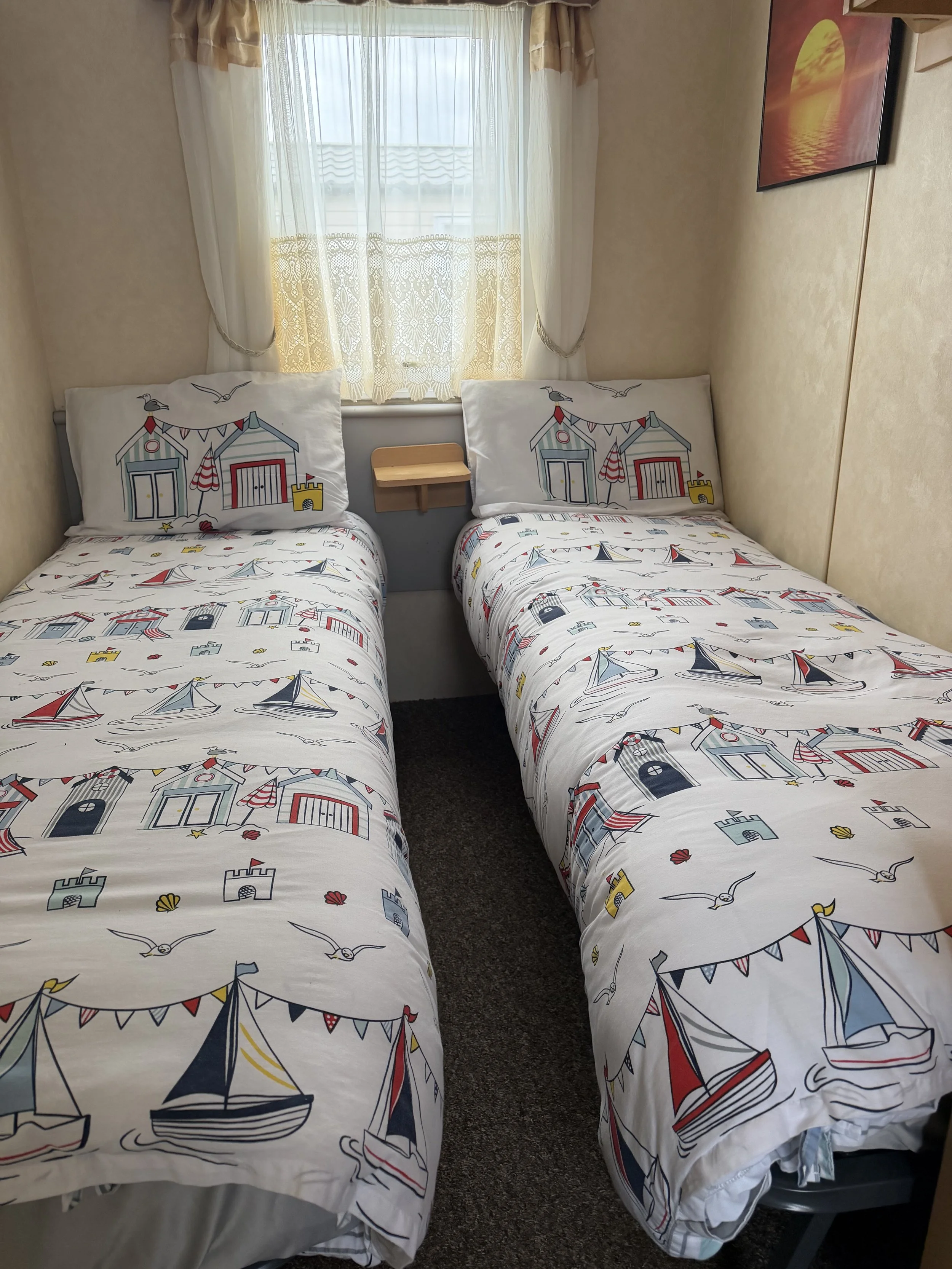 Two twin beds with nautical-themed bedspreads featuring sailboats and seaside elements, positioned side by side in a small bedroom. Each bed has pillows with drawings of beach huts. A window with curtains is behind the beds, and a painting of a sunse