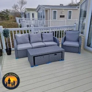 Outdoor patio setup with a gray wicker sofa, a matching armchair, and a rectangular storage ottoman, on a light-colored deck with a white fence and neighboring mobile homes in the background.