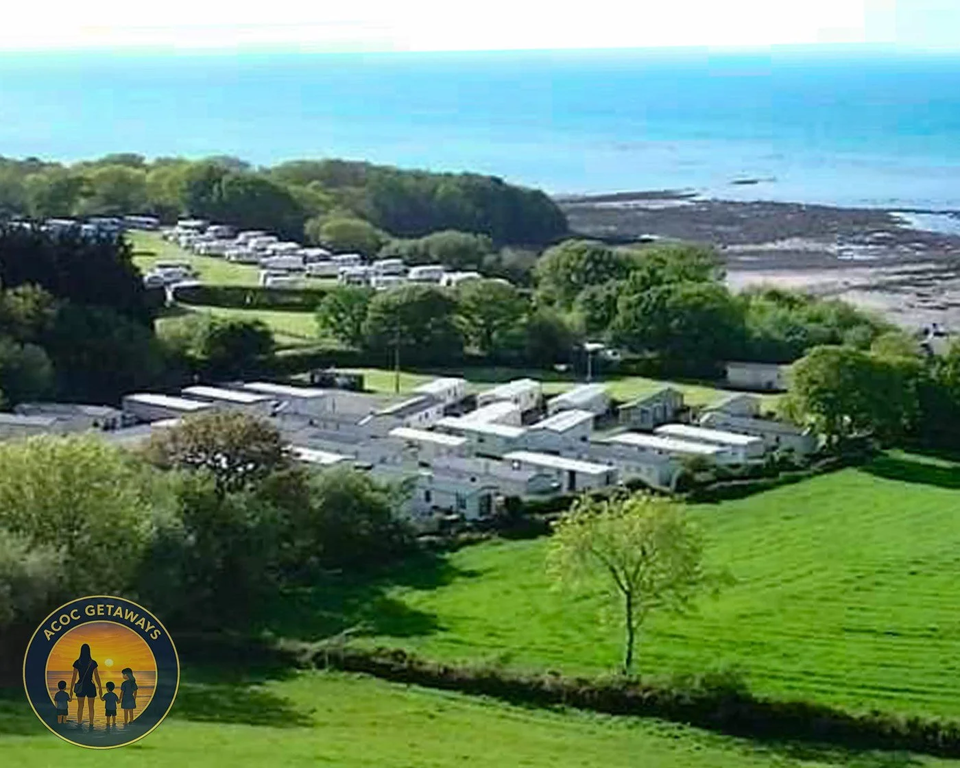 Aerial view of a coastal park with green lawns, trees, and white mobile homes, near the ocean with rocky shoreline.