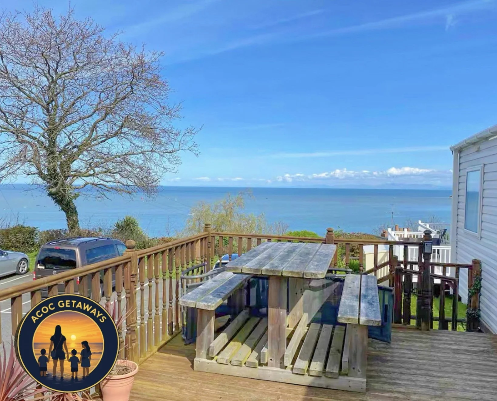 A backyard deck with outdoor furniture overlooking a large body of water under a blue sky with some clouds. There is a tree without leaves on the left and several cars parked below. The deck has a wooden railing with posts and a table with benches. P
