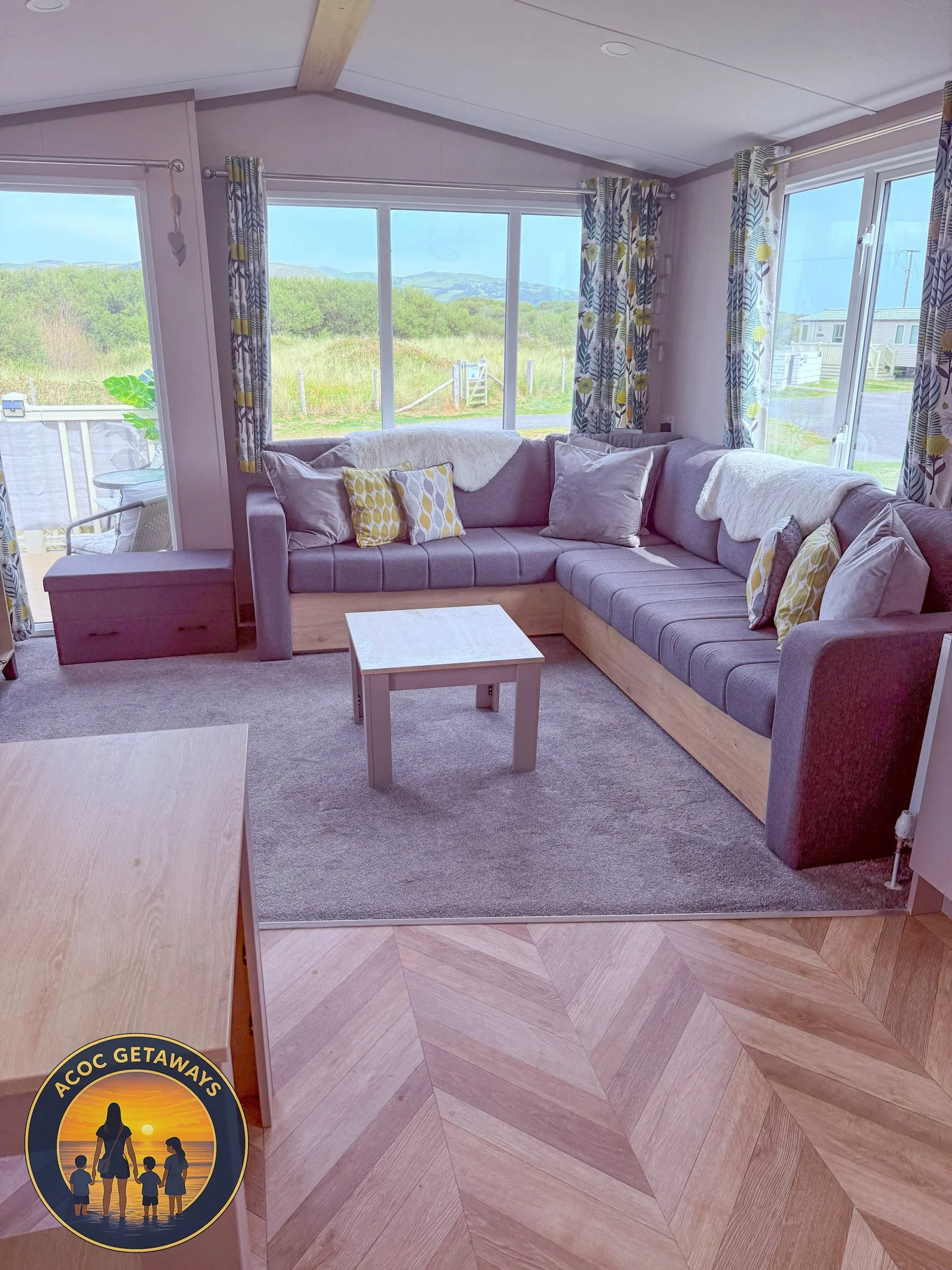 Cozy living room with a sectional sofa, decorative pillows, large windows with patterned curtains, a small coffee table, and a view of greenery outside.