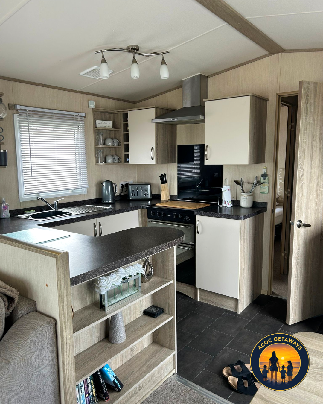 Kitchen with beige cabinets, black countertops, window with blinds, and various appliances including a stove, toaster, kettle, and utensils. Open shelving displays cups and glasses, and a hallway leading to a bedroom is visible.