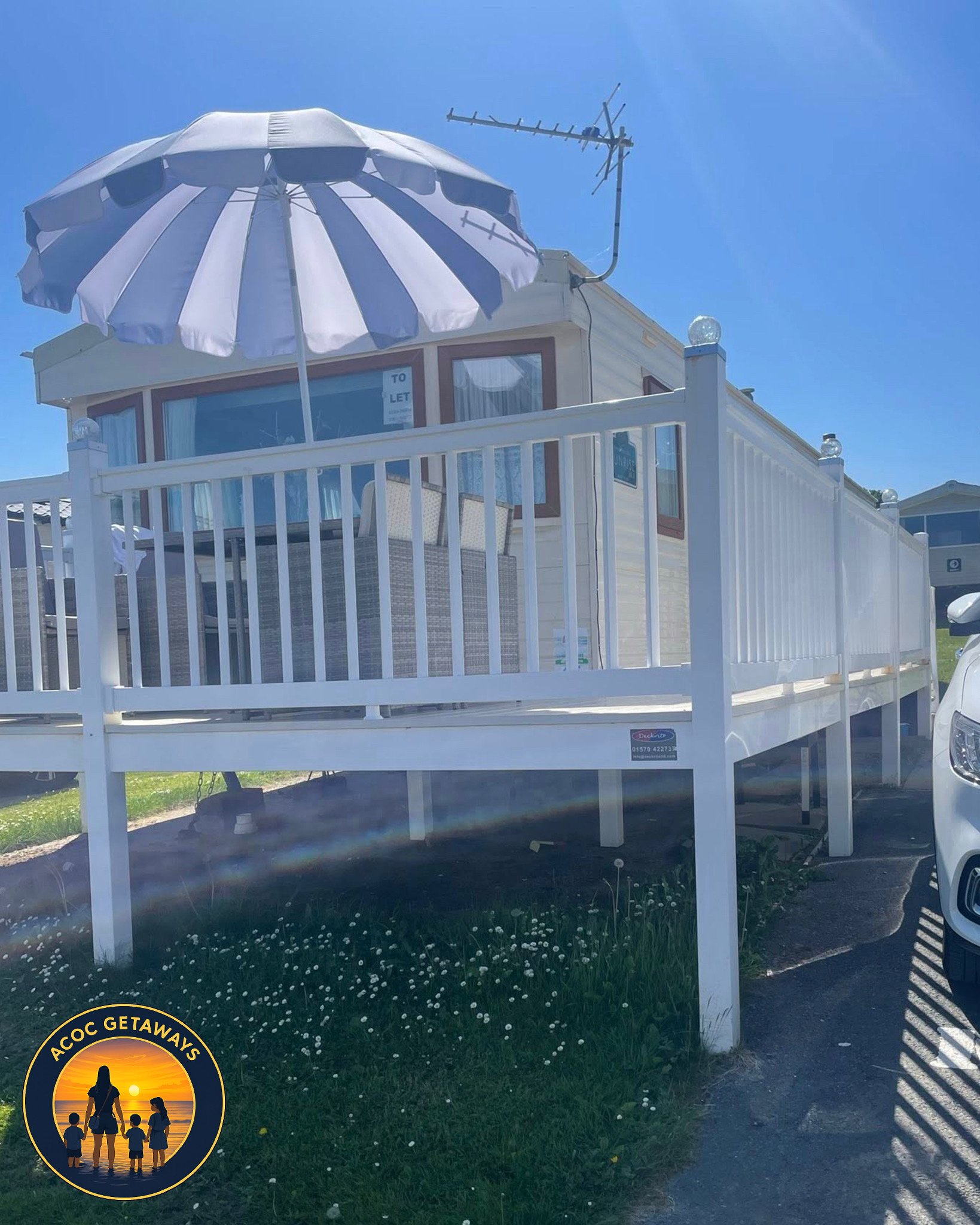 A small beach house with white railings, a large striped beach umbrella, and a TV antenna on the roof, set on a grassy area next to a parking lot with cars.
