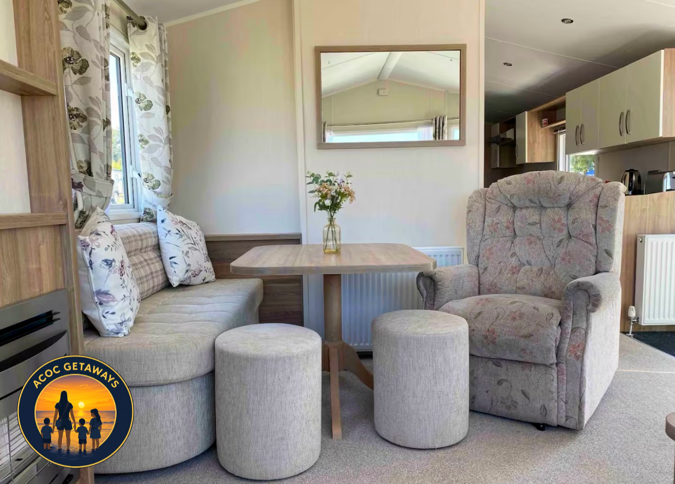 A cozy living area with a small wooden dining table, a floral-patterned armchair, a beige cushioned bench with pillows, two round ottomans, a mirror on the wall, a window with floral curtains, and a view of the kitchen area in the background.