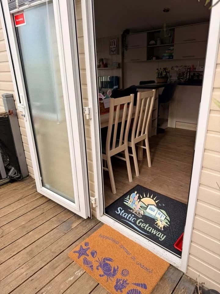 View of a house interior seen through an open glass door from a wooden outside deck. Inside, there is a dining table with four chairs, and a countertop with various kitchen items. A black welcome mat with a sunset, house, dog, and the words "Static G