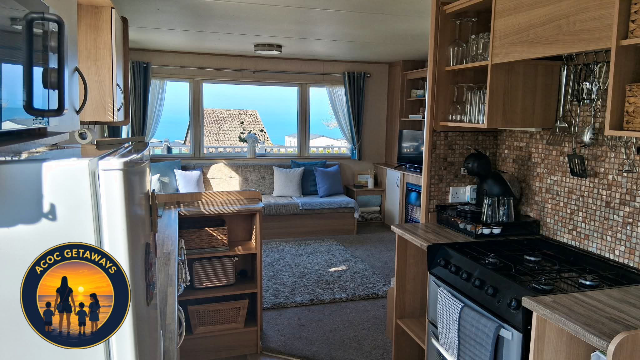 Inside a cozy beachfront cottage with a kitchen and living area, featuring wooden cabinets, a stove, a countertop with coffee maker, and a large window showing the ocean and rooftops outside.