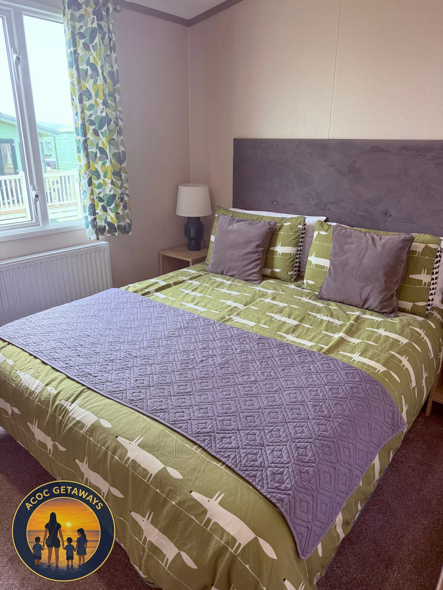 A bedroom with a bed that has green and white patterned bedding, two gray pillows, and a purple quilt at the foot of the bed. There is a window with patterned curtains and a nightstand with a black lamp and a white lampshade.