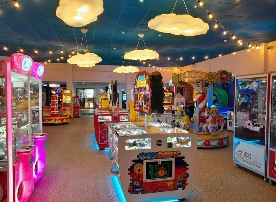 Inside an arcade with various claw machines, colorful decorations, and ceiling lights shaped like clouds and string lights.
