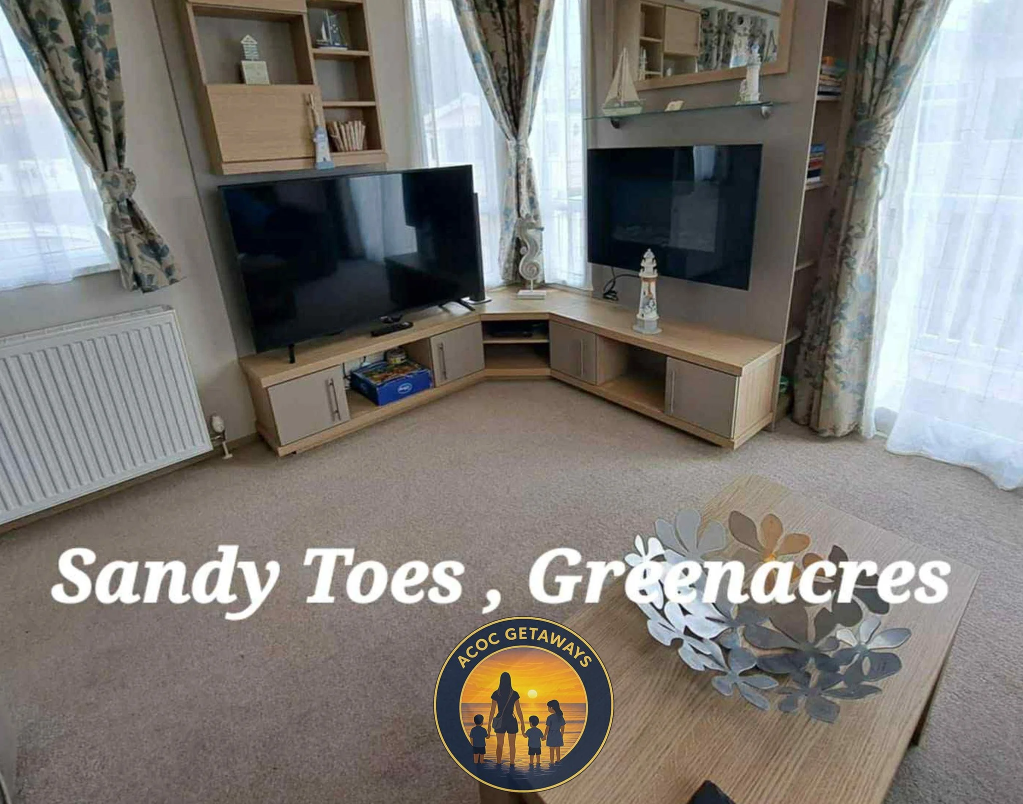 Living room with beige carpeting, two large flat-screen TVs on a corner TV stand, with decorative model ships on shelves, floral curtains, and a round wooden coffee table with a metal decorative centerpiece.