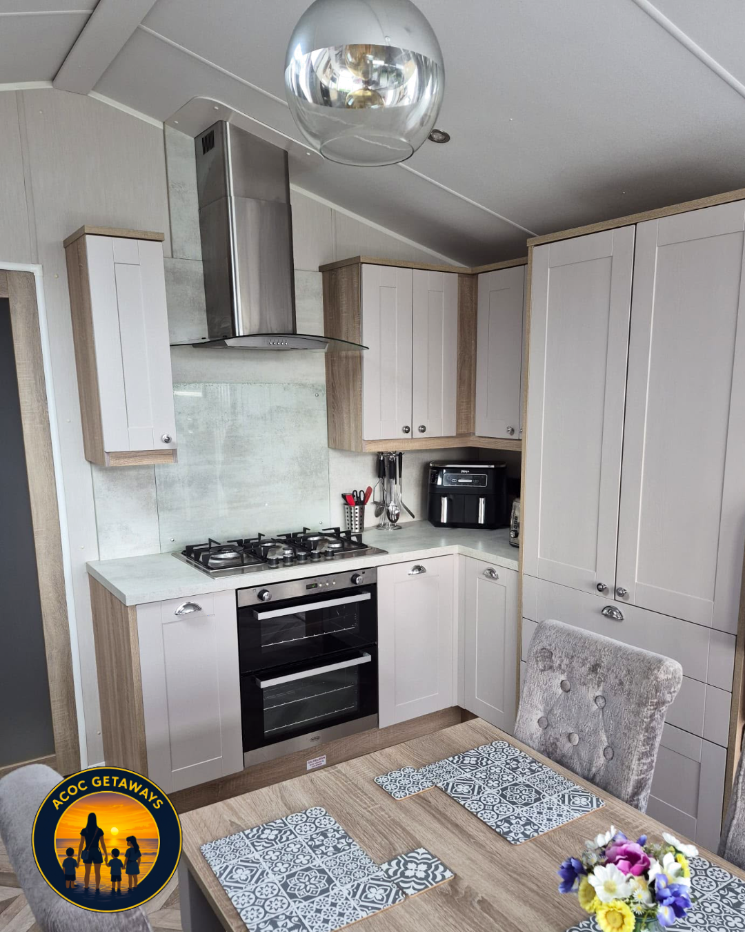 Modern kitchen with beige cabinets, stainless steel stove and hood, black air fryer, and a dining table with patterned placemats and a flower centerpiece.