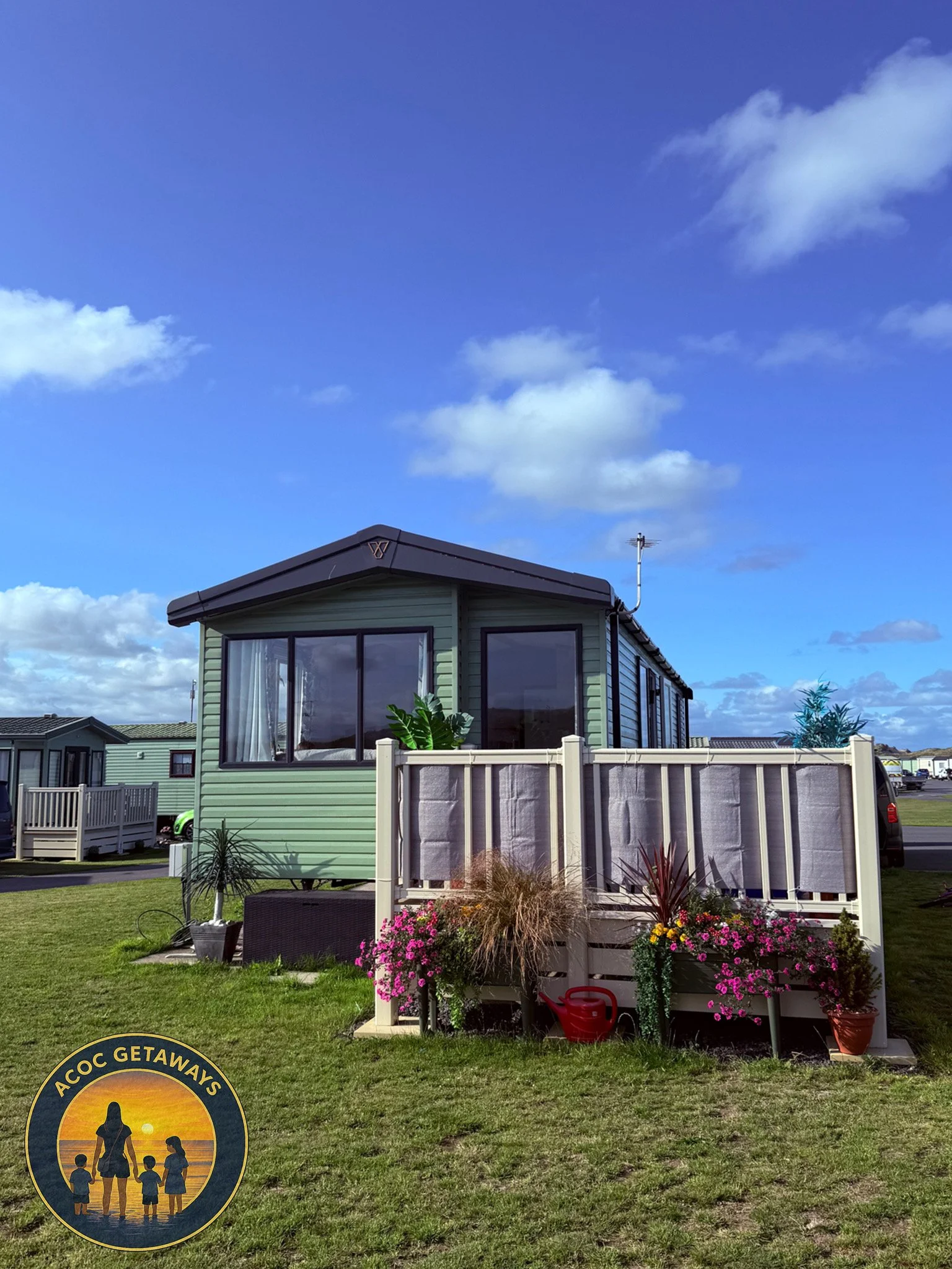 A small, light green mobile home with a front porch decorated with pink flowers and potted plants, set on a grassy area under a partly cloudy blue sky.