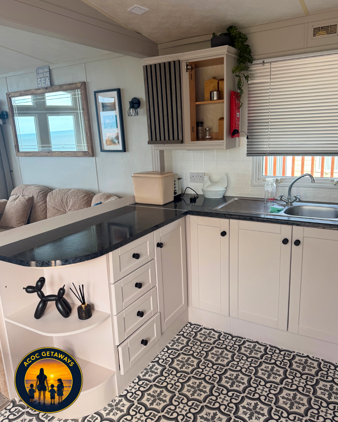 Kitchen with white cabinets, black countertops, a window with blinds, and a sink. Decor includes a framed photo, a wall lamp, a small open cabinet, and decorative items. Patterned black and white floor tiles, a beige couch with throw pillows visible 