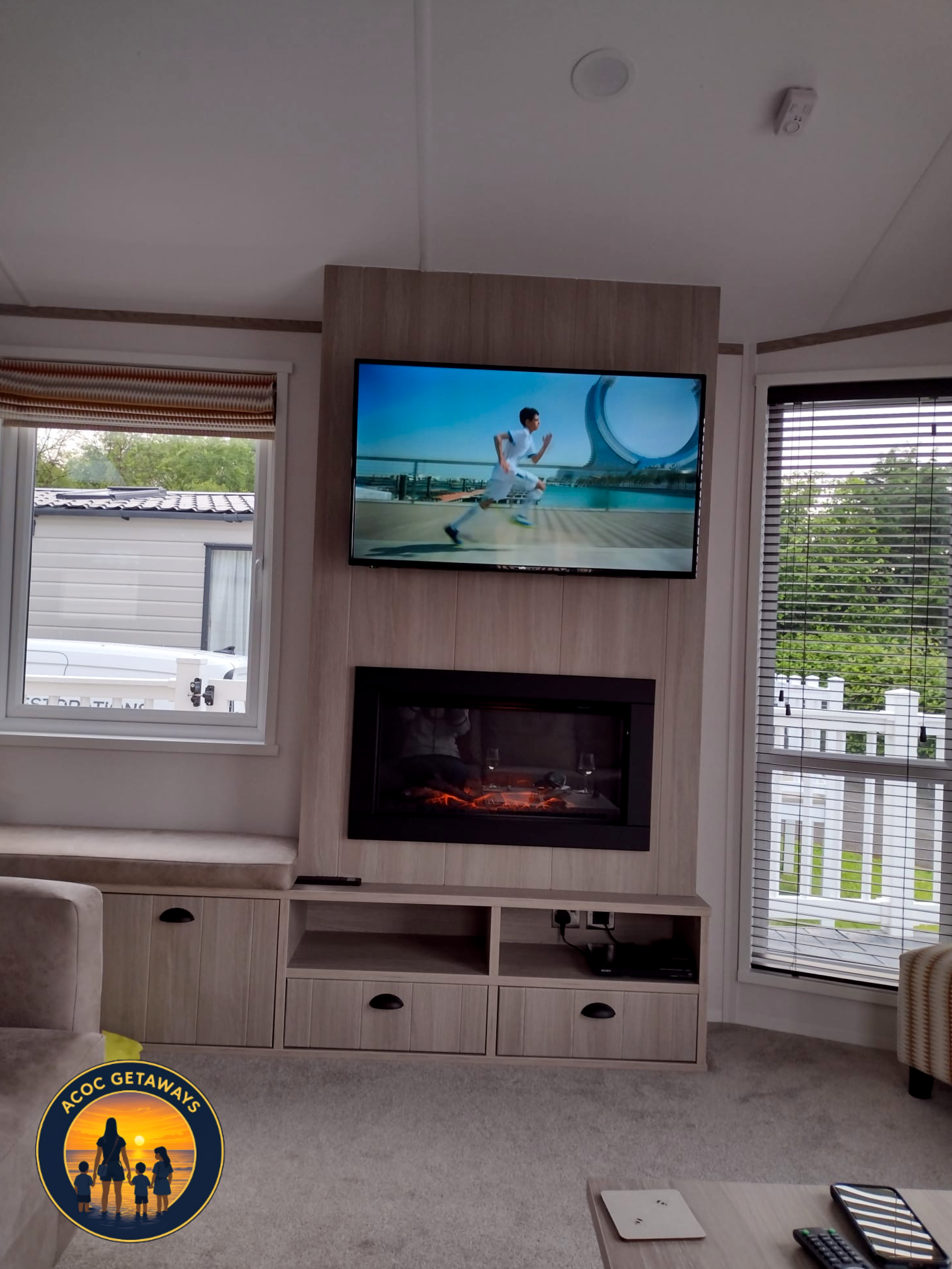 Living room with beige carpet, light-colored wooden entertainment center, television displaying a person running outdoors, electric fireplace below TV, large windows with blinds showing outside greenery, beige armchair partially visible, and a TV rem