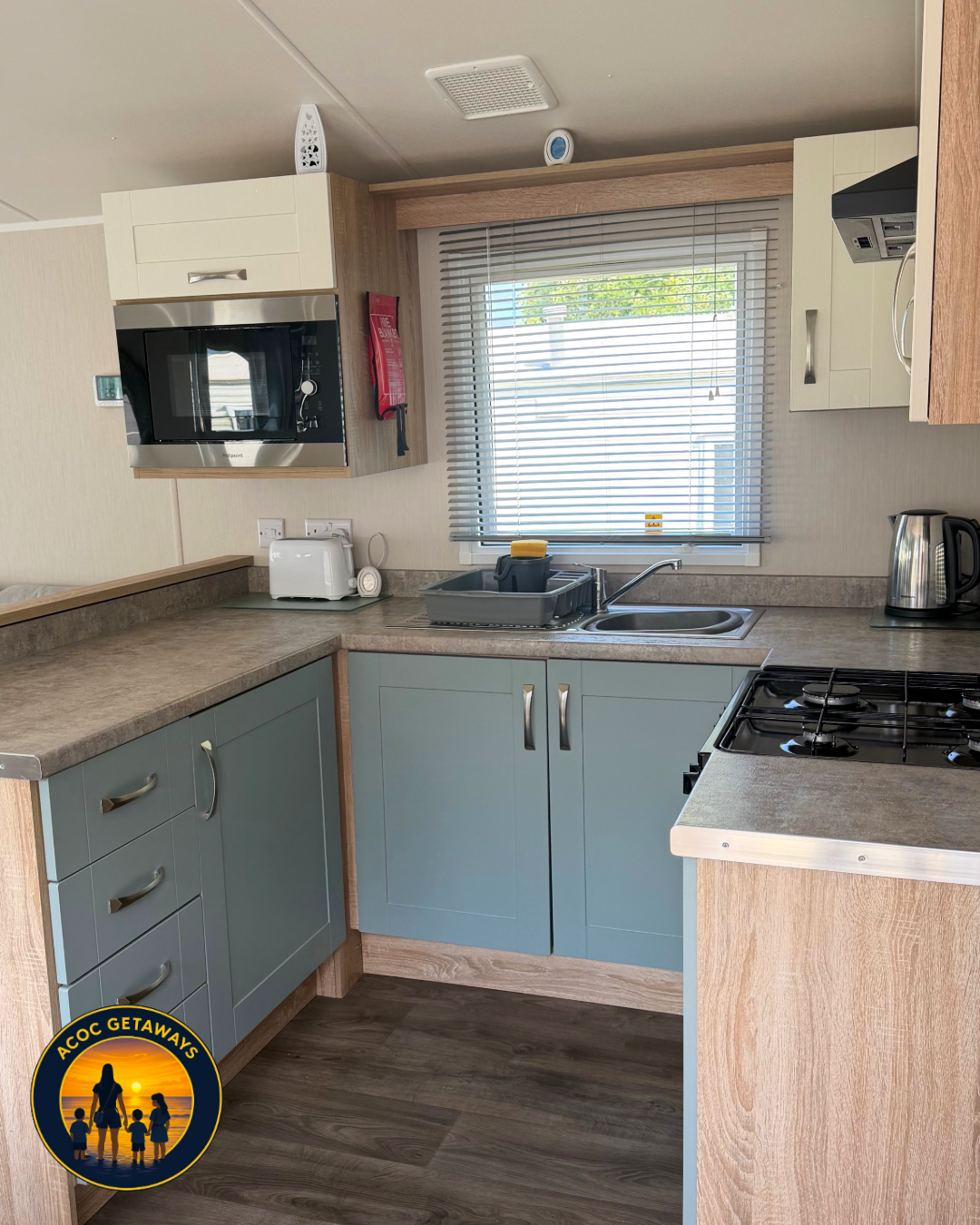 Kitchen with light blue cabinets, a window with blinds, a gray countertop, a sink with dishes, a microwave, toaster, kettle, and stove.