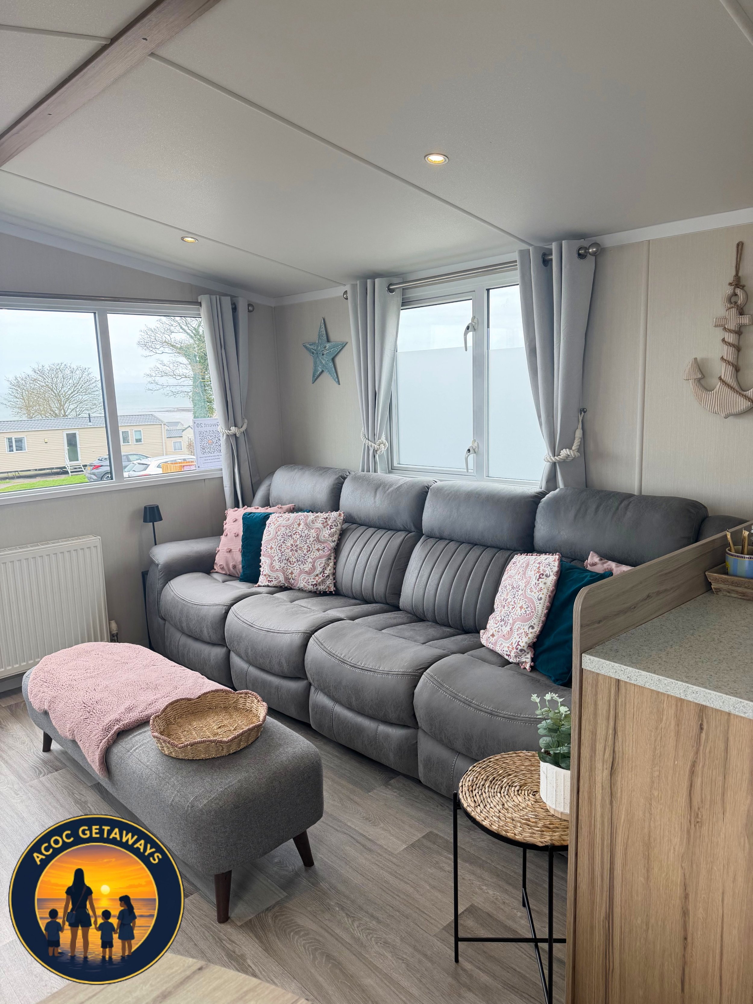 Living room with a gray sofa, decorated with pink, white, and teal pillows. A pink blanket and a woven basket are on an ottoman in front of the sofa. Large windows with white curtains and a view of trees and mobile homes outside. A small black side t