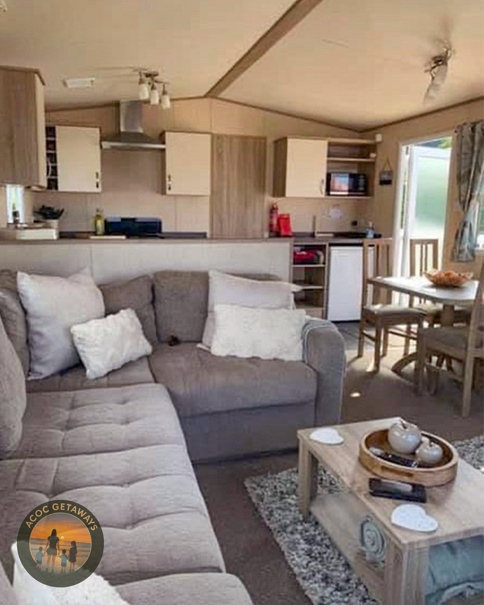 Living room with beige sofas, a wooden coffee table, a dining table with chairs, and an open kitchen with cabinets, microwave, and small refrigerator, with natural light coming through a sliding door.