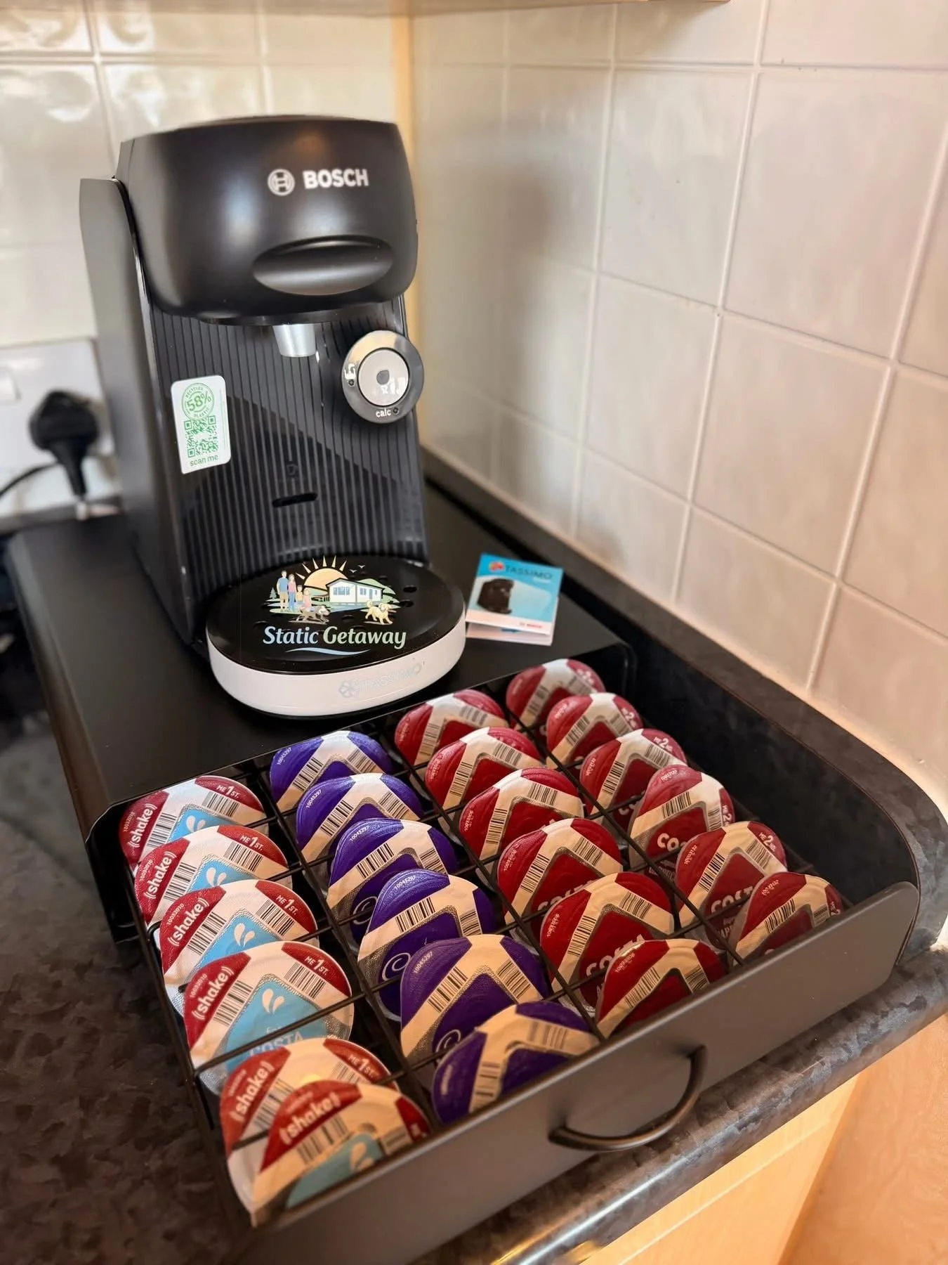 A coffee station featuring a Bosch coffee machine, a tray of flavored coffee creamers, and coffee pods on a black countertop against a tiled wall.