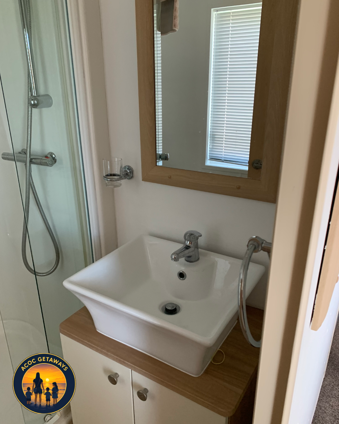 Small bathroom with a white sink, a mirror with a wooden frame, a window with blinds, and a shower with a glass door.