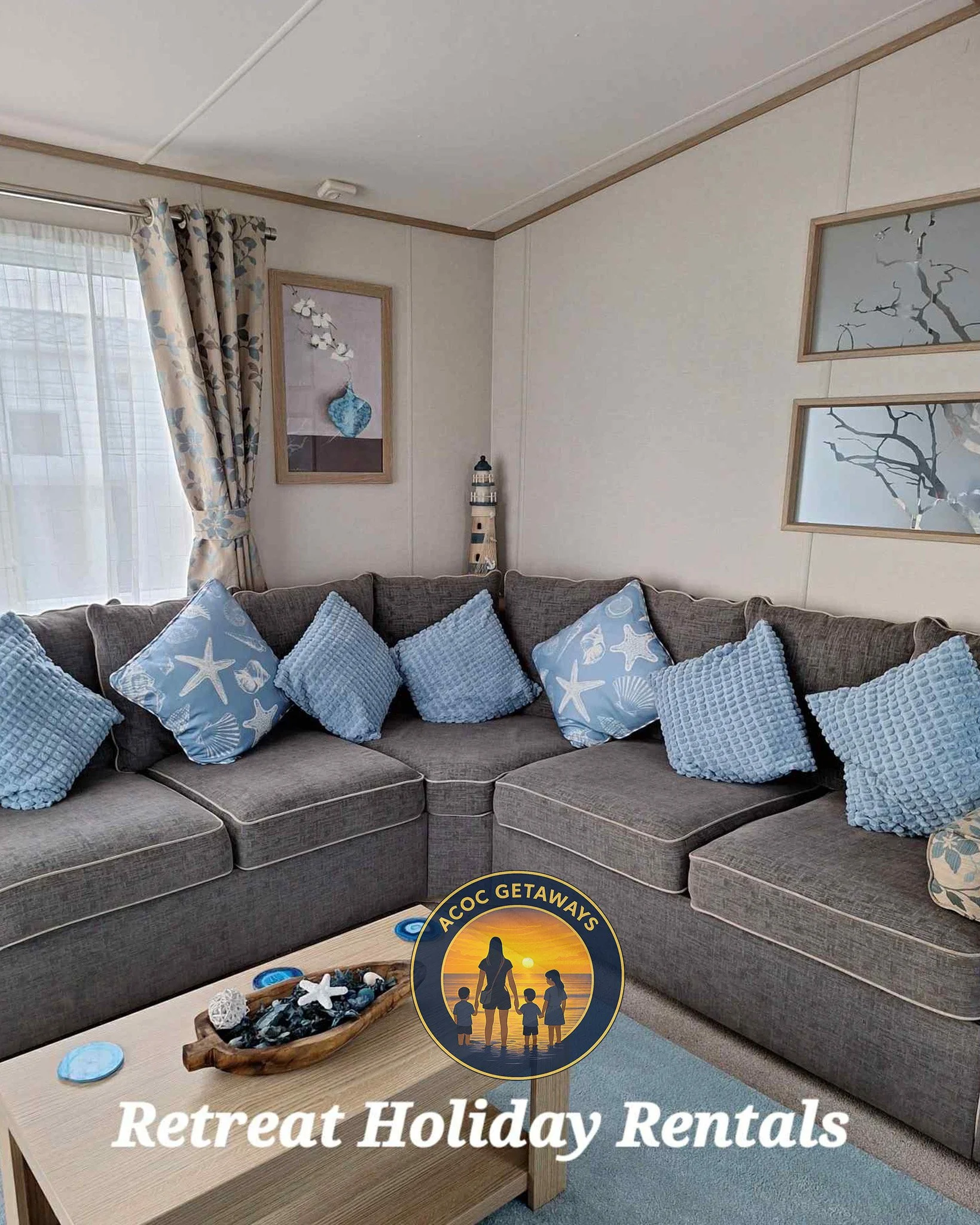A cozy living room with a gray sectional sofa decorated with blue throw pillows and a wooden coffee table with surf-themed decorations. There are framed ocean and nature-themed artwork on the walls, a window with patterned curtains, and a room logo for 'ACOC Getaways Retreat Holiday Rentals' at the bottom.