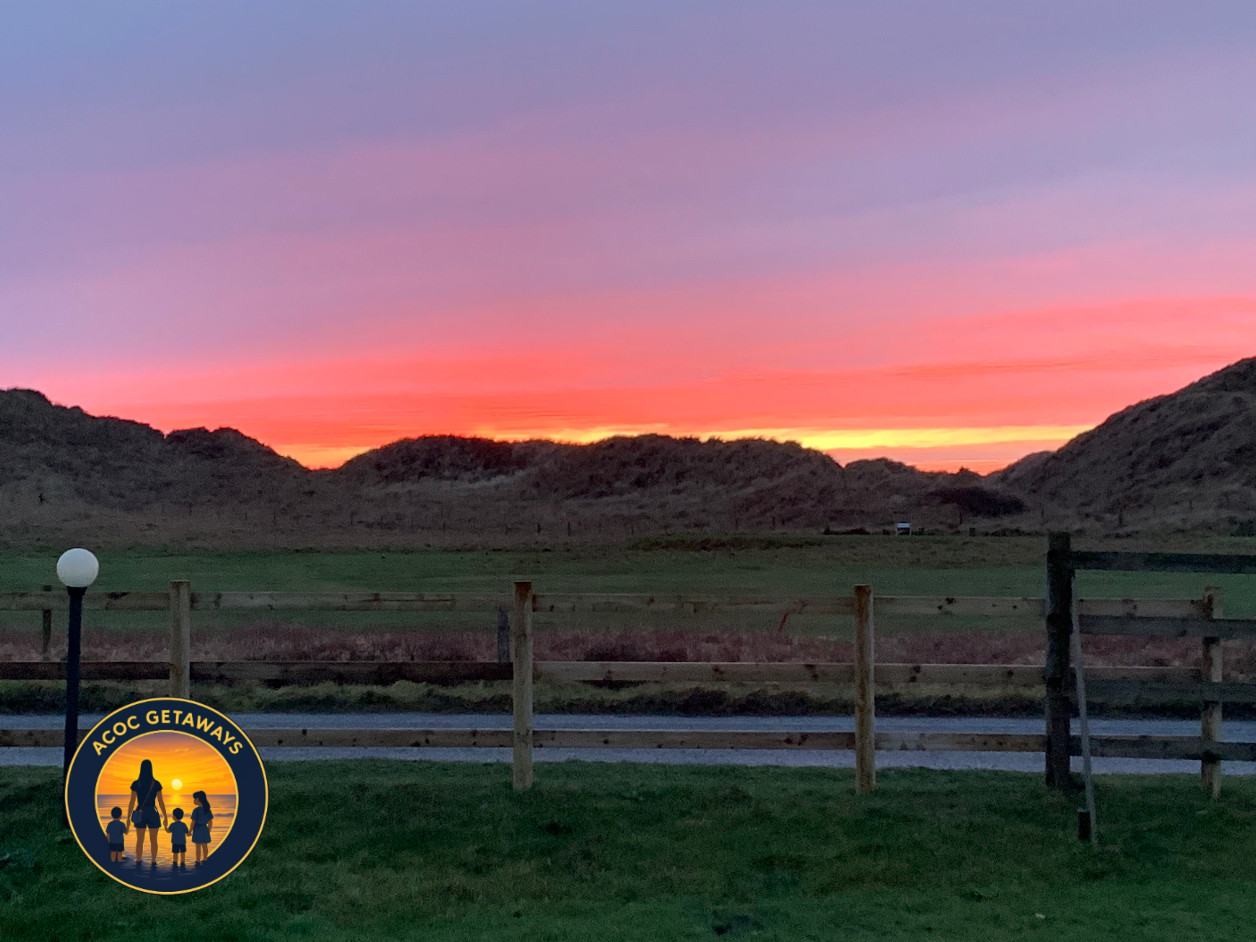 Sunset over a grassy field with rolling hills in the distance, wooden fence, and a lamppost in the foreground. In the lower left corner is a logo with a silhouette of a woman and children at sunset with the text 'ACOC GETAWAYS'.