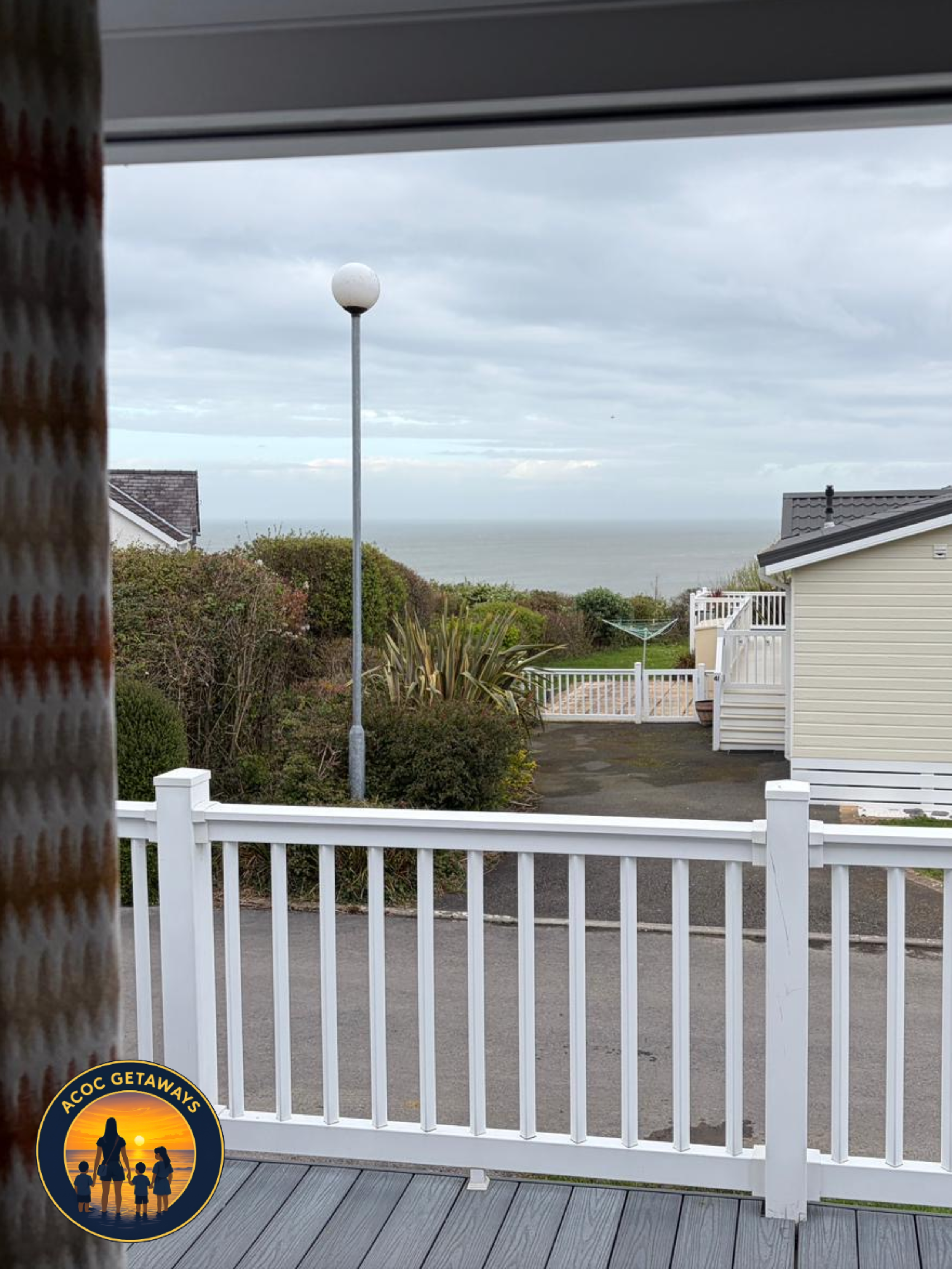 View from a porch looking out over a fence, street, bushes, houses, and ocean with cloudy sky in the background.