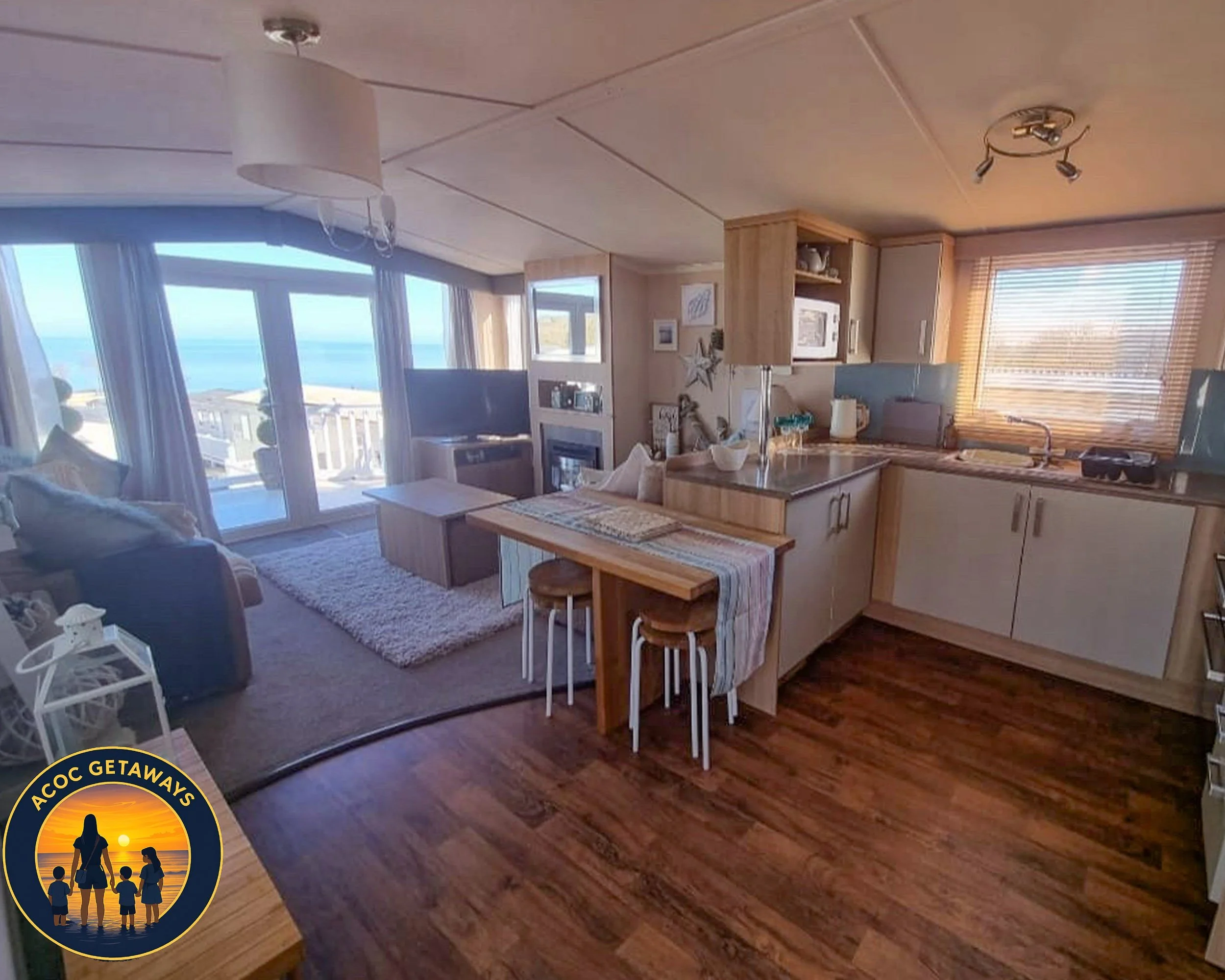 Open-concept living room and kitchen with large windows showing a balcony and ocean view, wooden floors, beige and light-colored furniture, small dining table, TV, and kitchen appliances.