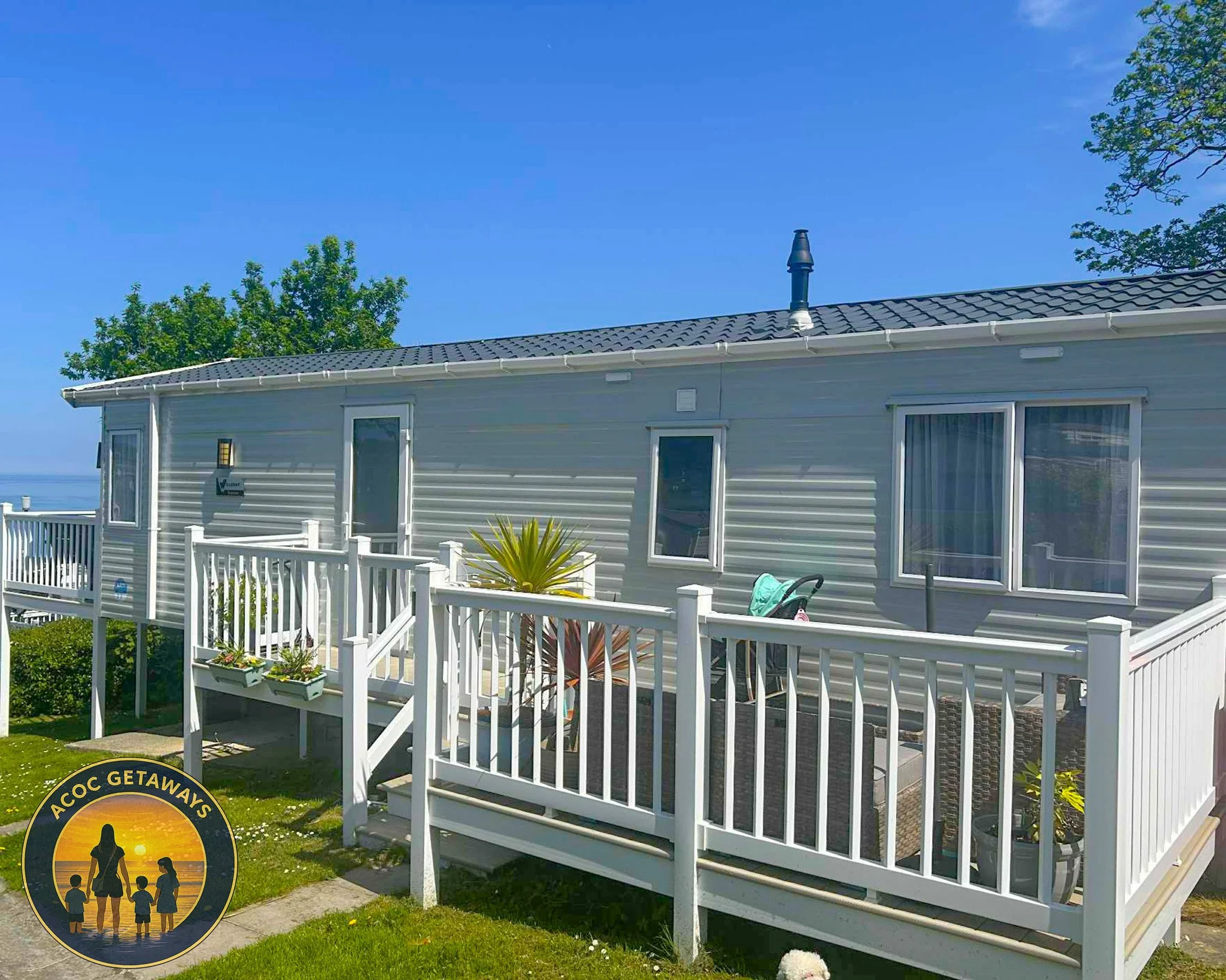 A light gray manufactured home with a white porch and railing, three windows, potted plants, and patio furniture, set against a clear blue sky with trees in the background.