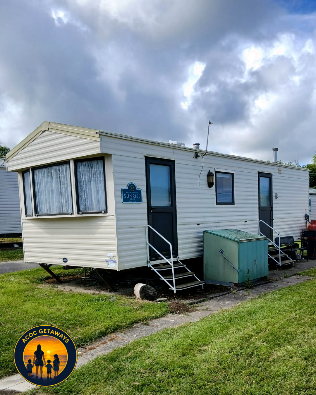 White mobile home with black window frames, steps leading to doors, green storage container outside, blue sky with clouds.