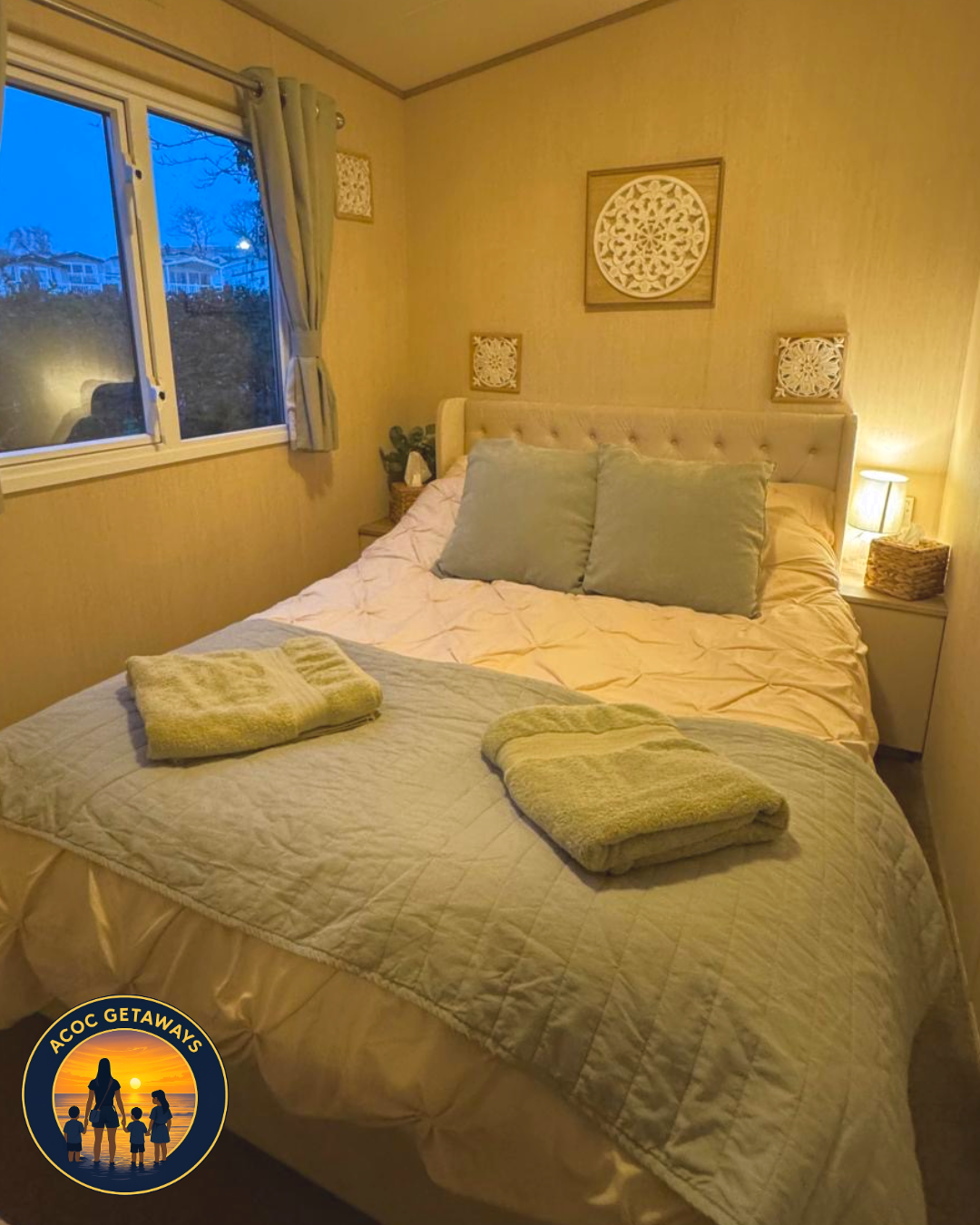 A cozy bedroom with a bed that has a beige tufted headboard, light-colored bedding, and two large pillows. Two neatly folded towels are placed on a quilted blanket at the foot of the bed. There are nightstands on each side, each with a small lamp. Th