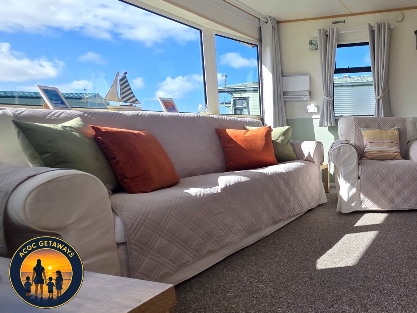 Living room with a large white sofa decorated with green and orange throw pillows, a matching armchair with a beige cushion, a small wooden side table, large windows with white curtains, and a view of a blue sky with clouds and nautical decorations o