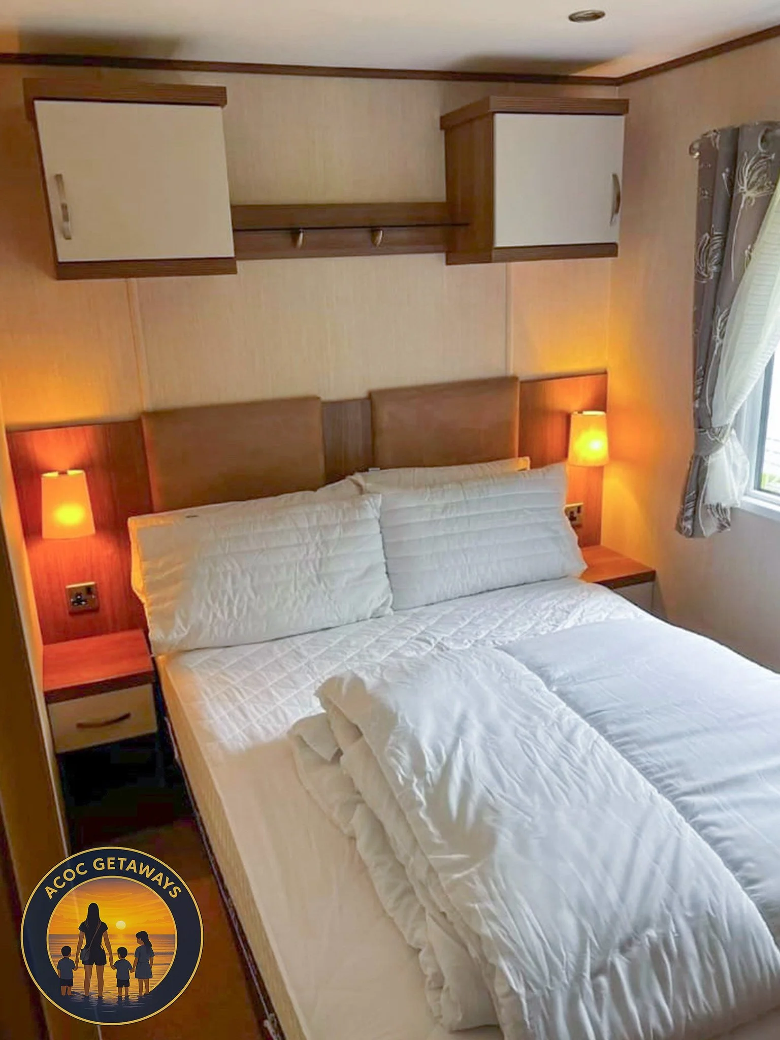 A cozy hotel bedroom with a neatly made bed, pillows, and a white comforter. There are two small bedside tables with orange lamps, a window with floral curtains, and wooden wall-mounted cabinets above the bed.