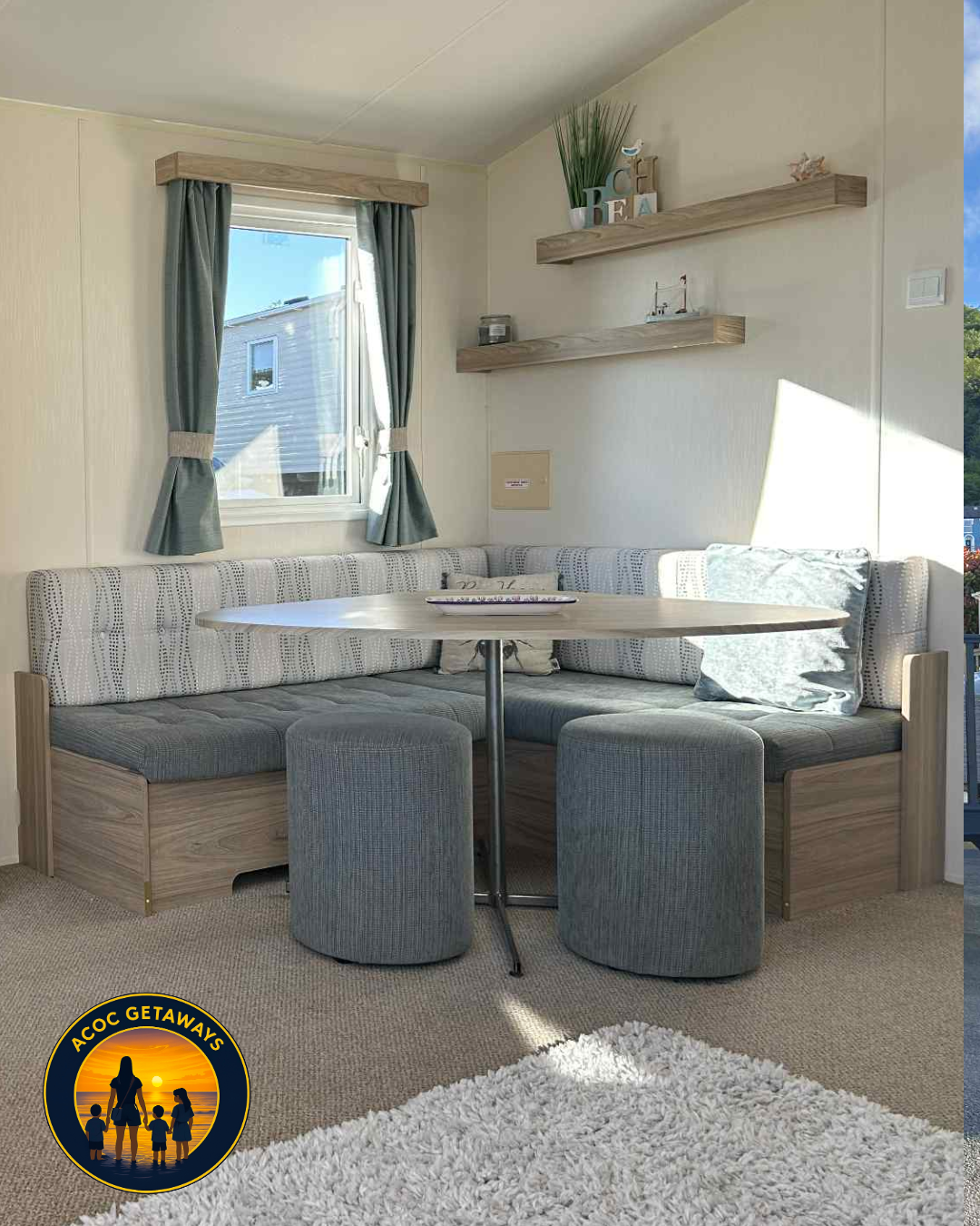 Beach-themed living room with window, green curtains, corner cushioned seating, small round table, and decorative shelves with plants and art.