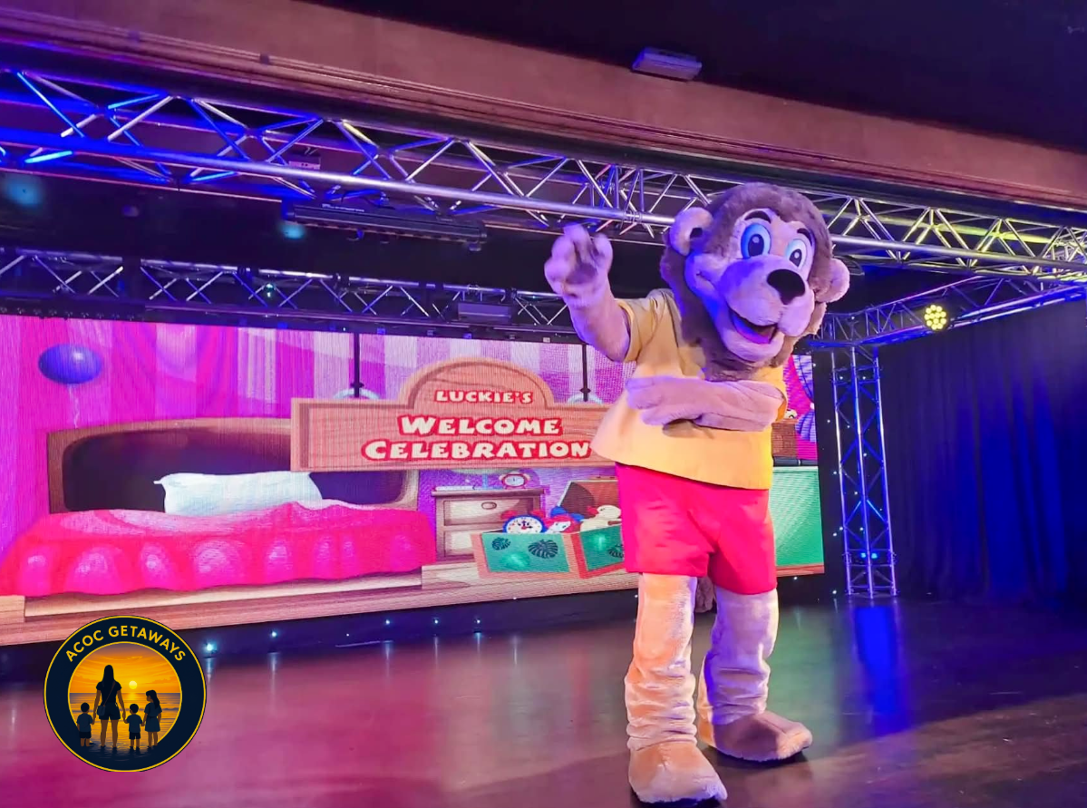 Costumed lion character on stage with a digital backdrop reading "Luckie's Welcome Celebration."