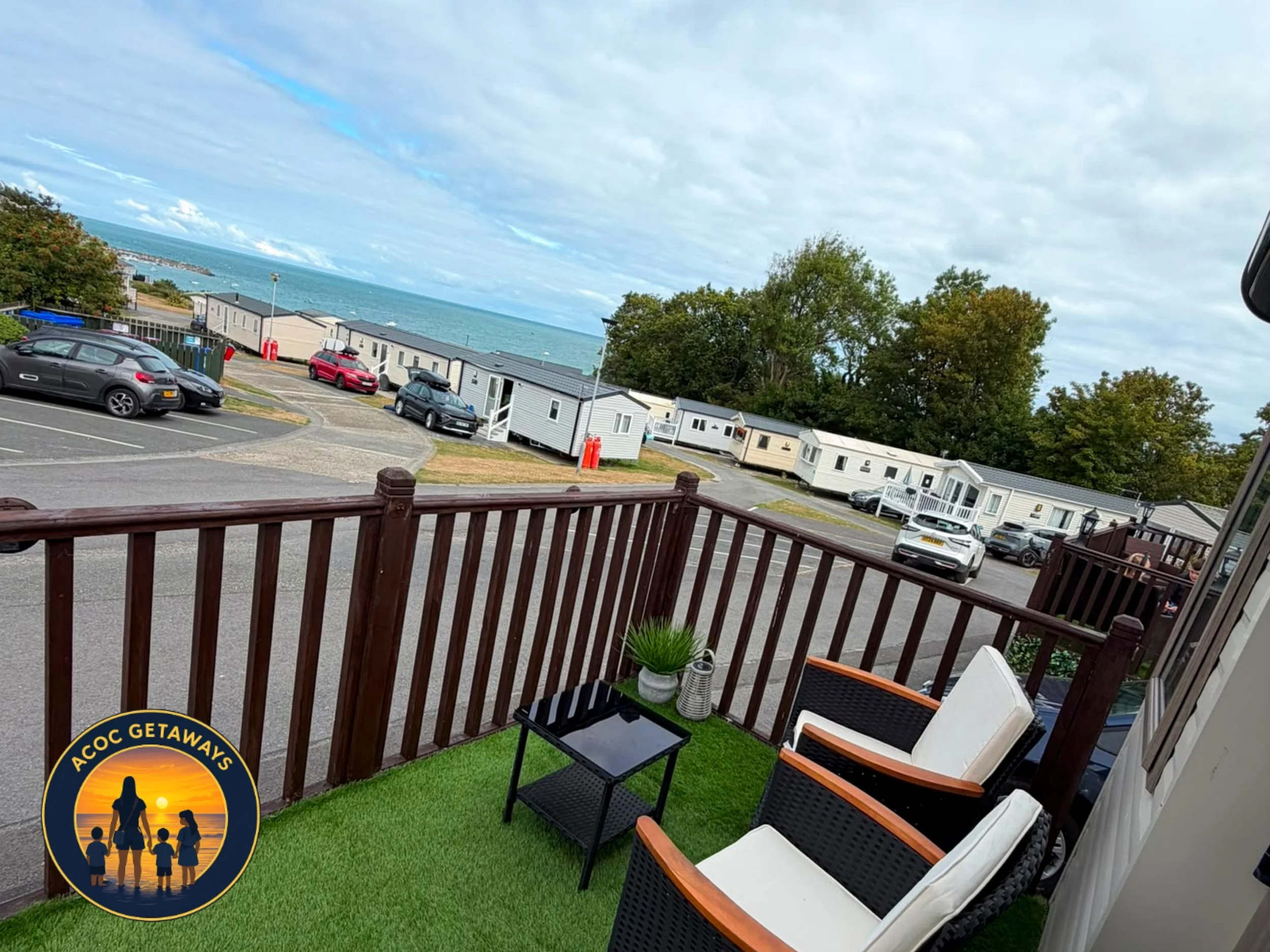 View from a balcony showing a grassy outdoor area with chairs and a small table, a parking lot with cars, white mobile homes, trees, and the ocean in the background under a cloudy sky.