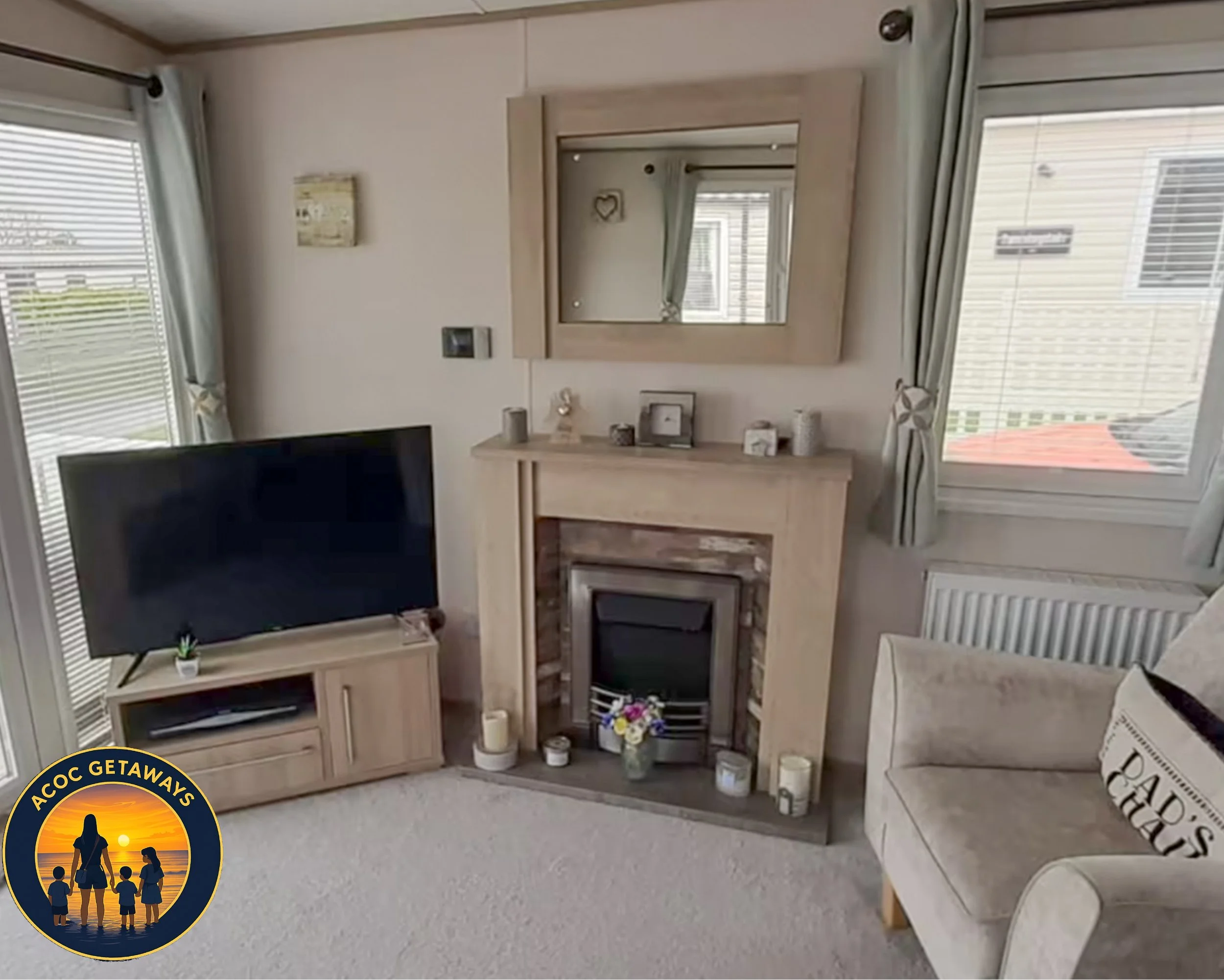 Living room with a beige fireplace, a large flat-screen TV on a wooden stand, a beige armchair with a decorative pillow, and windows with curtains letting in natural light.