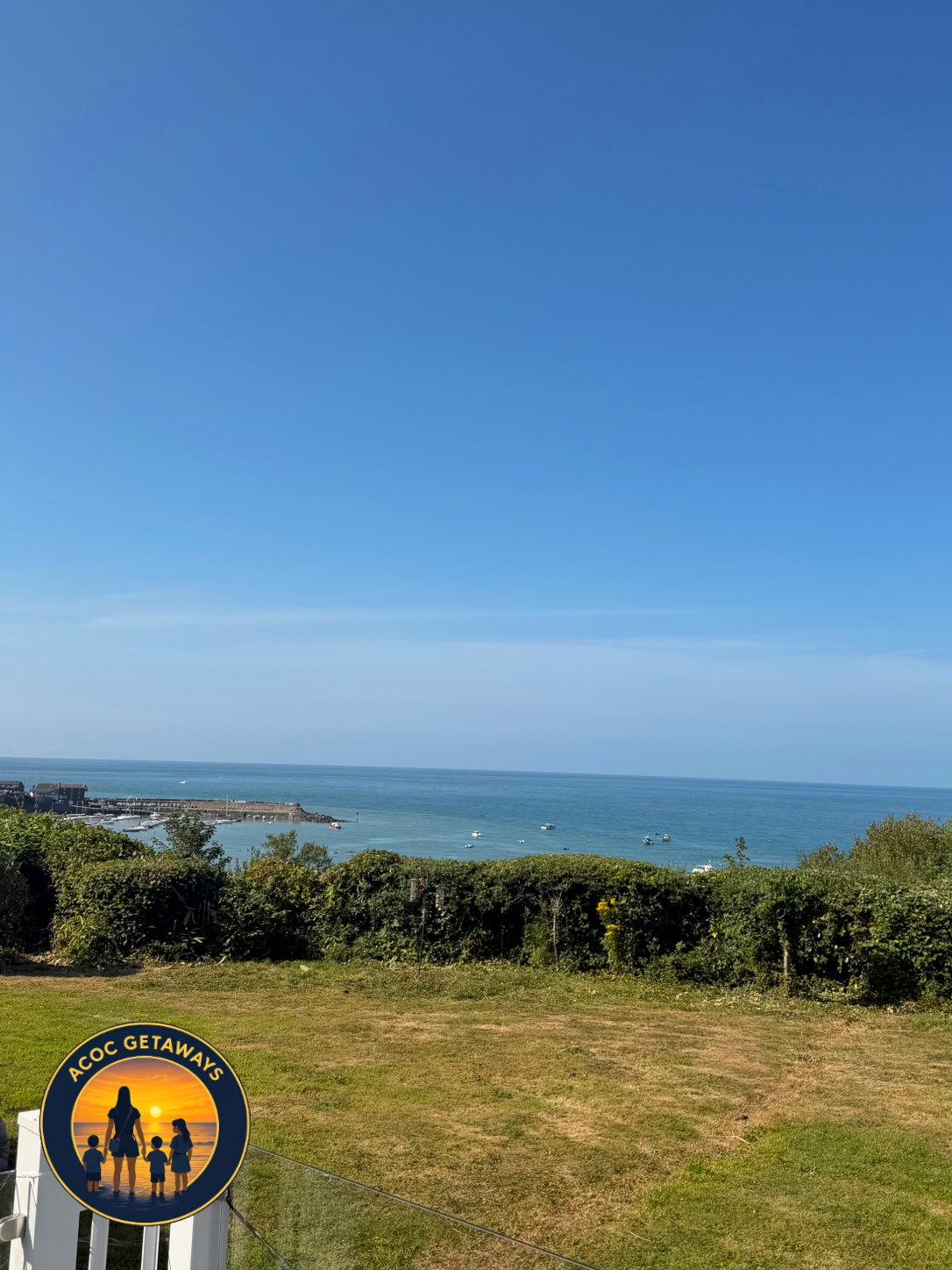 View of a blue sky and ocean from a grassy lawn with bushes in the foreground. There are small boats on the water and a coastline with buildings in the distance. A logo in the bottom left corner shows a silhouette family with sunset colors and the te