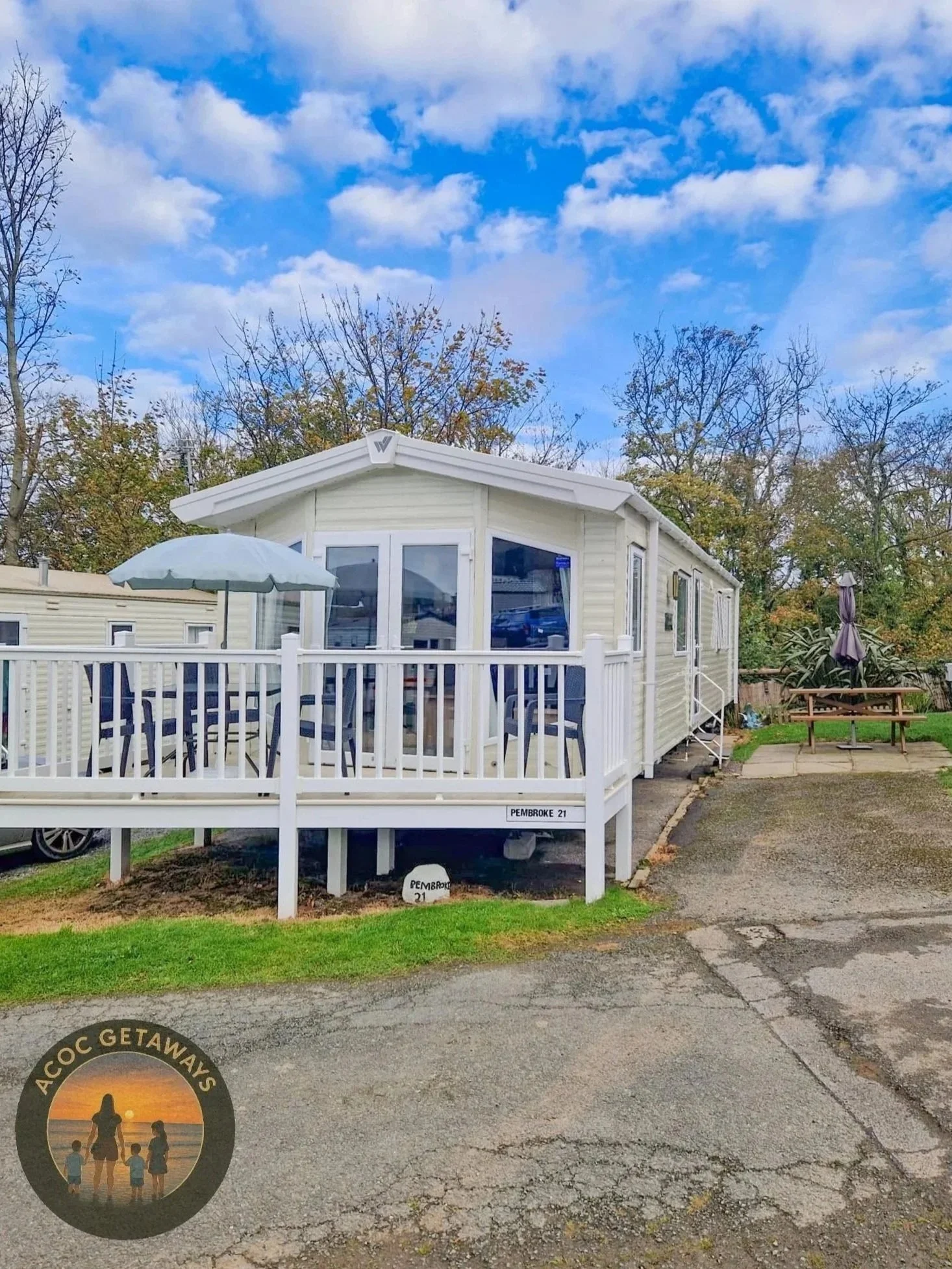 A white mobile home or caravan with a front porch and railing, set on a paved lot with surrounding grass and trees with autumn foliage, under a partly cloudy sky.