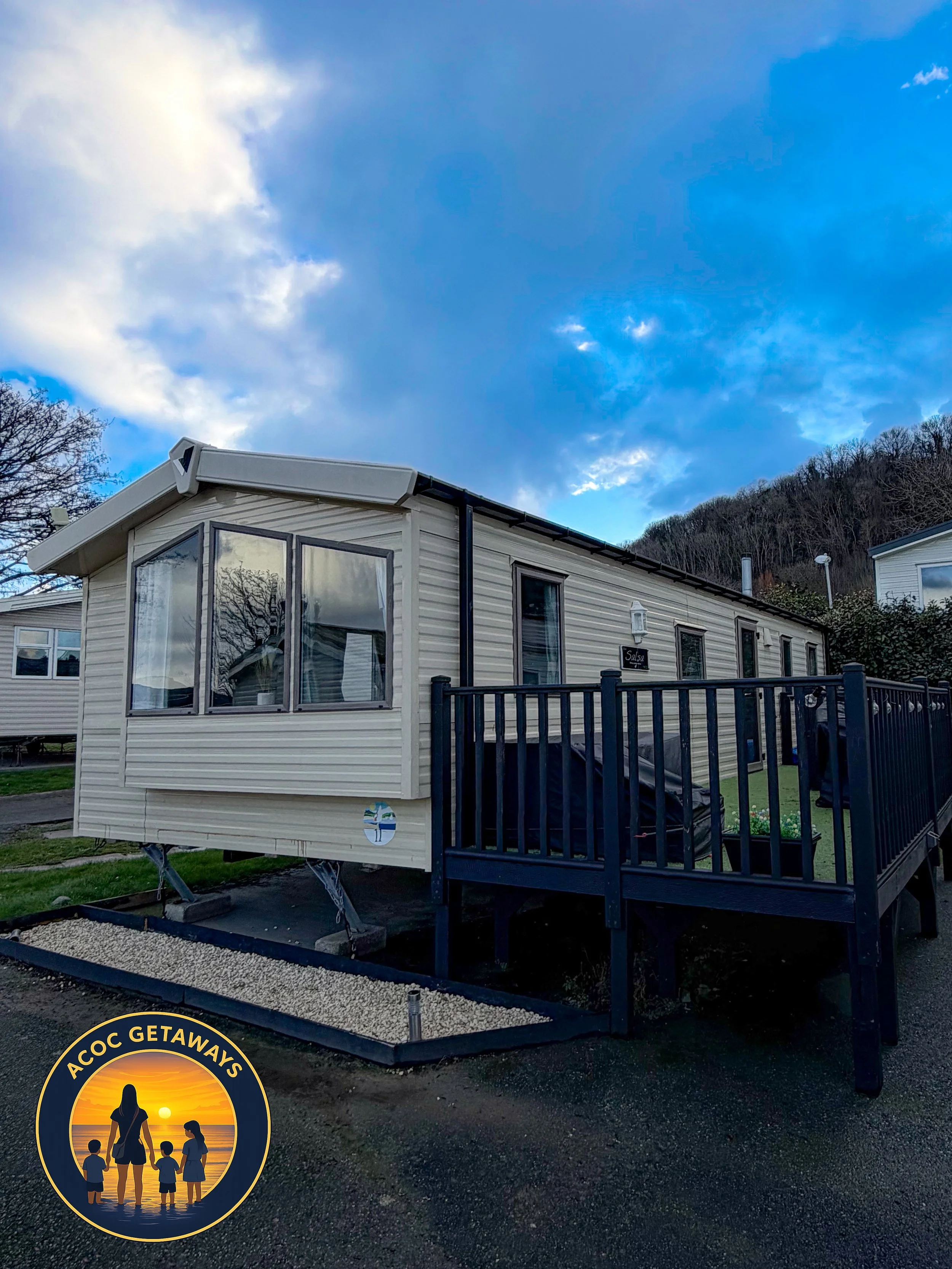 A beige mobile home with large windows and a small deck, situated in a park with a surrounding fence, under a partly cloudy sky, with a wooded hill in the background.