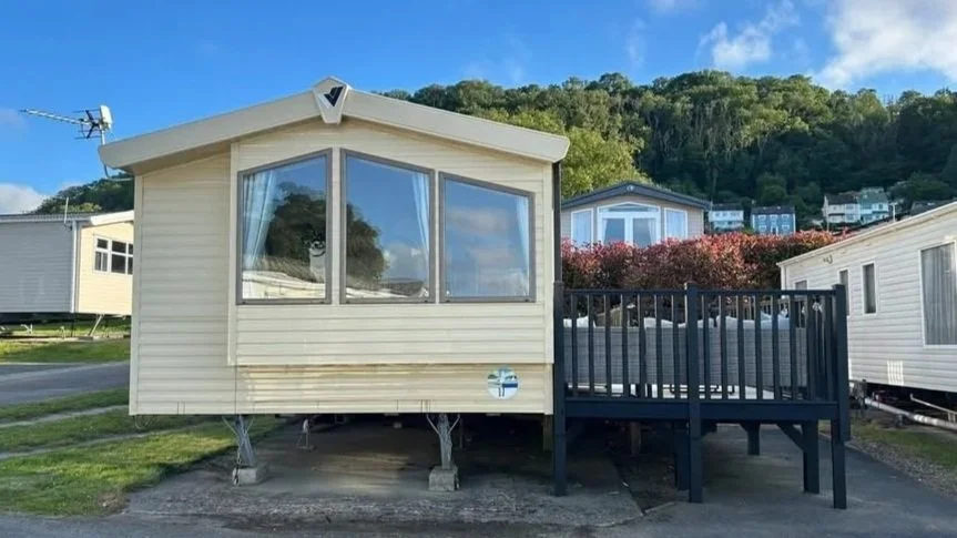A beige mobile home with large front windows and a black wooden porch, set on a gravel lot with other similar homes and a hillside in the background.
