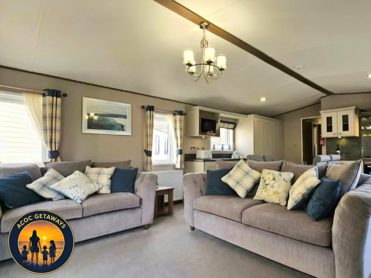 Living room with two beige sofas with patterned cushions, a window with checkered curtains, a small wooden side table, a framed picture of a landscape, a kitchen area with cabinets and a television, and a chandelier hanging from the ceiling.