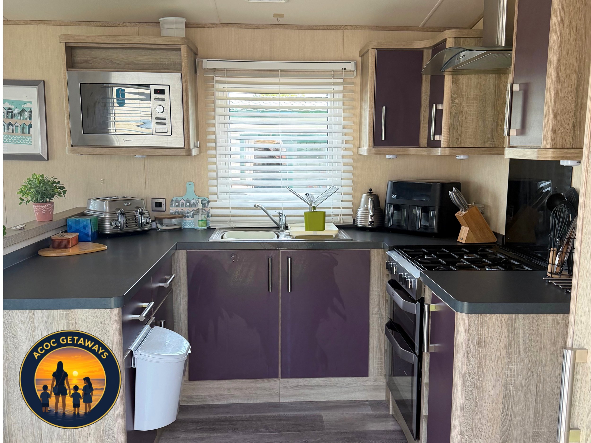 A compact kitchen with wooden cabinets, a window with blinds, a microwave above the counter, a toaster, a small plant, dish soap, a dish drying rack, a sink, and a stove with utensils. There is also an ACOT Getaways sticker at the bottom left corner.