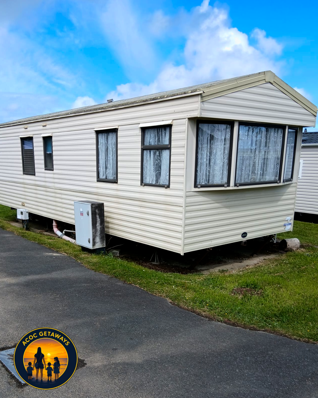 A beige mobile home with black window frames and lace curtains, situated on a grassy patch next to a paved road, under a partly cloudy blue sky.