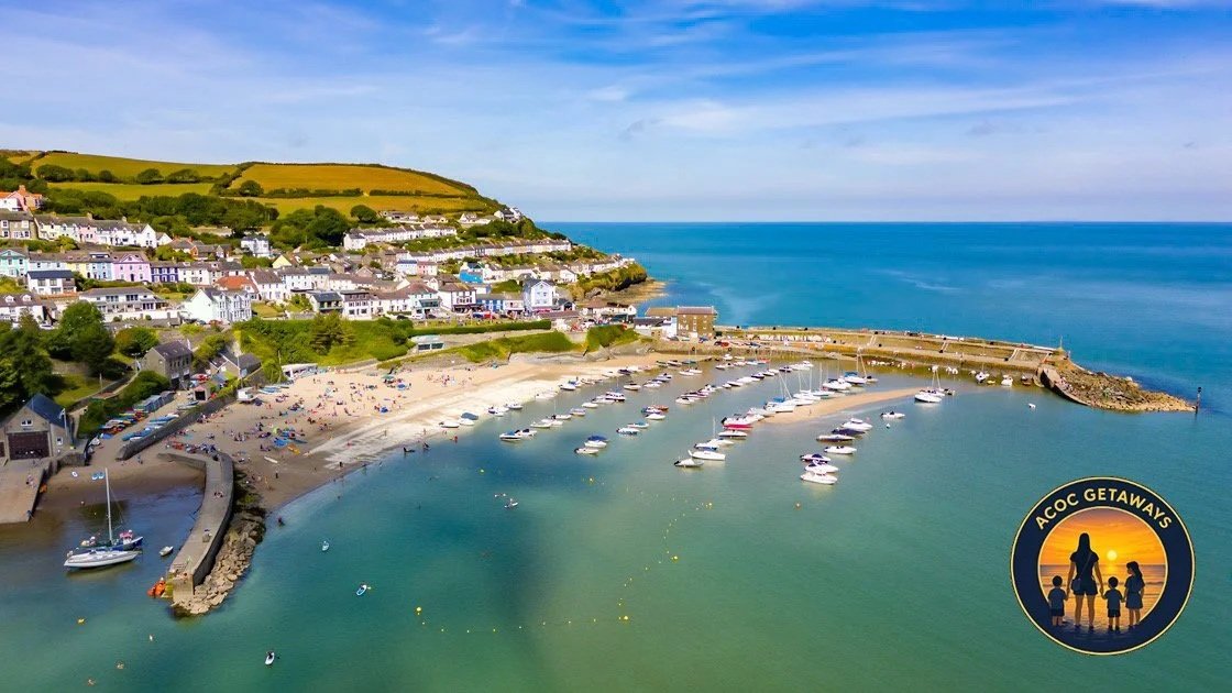 Aerial view of a harbor with boats, a sandy beach, and colorful houses on a hillside overlooking the ocean.