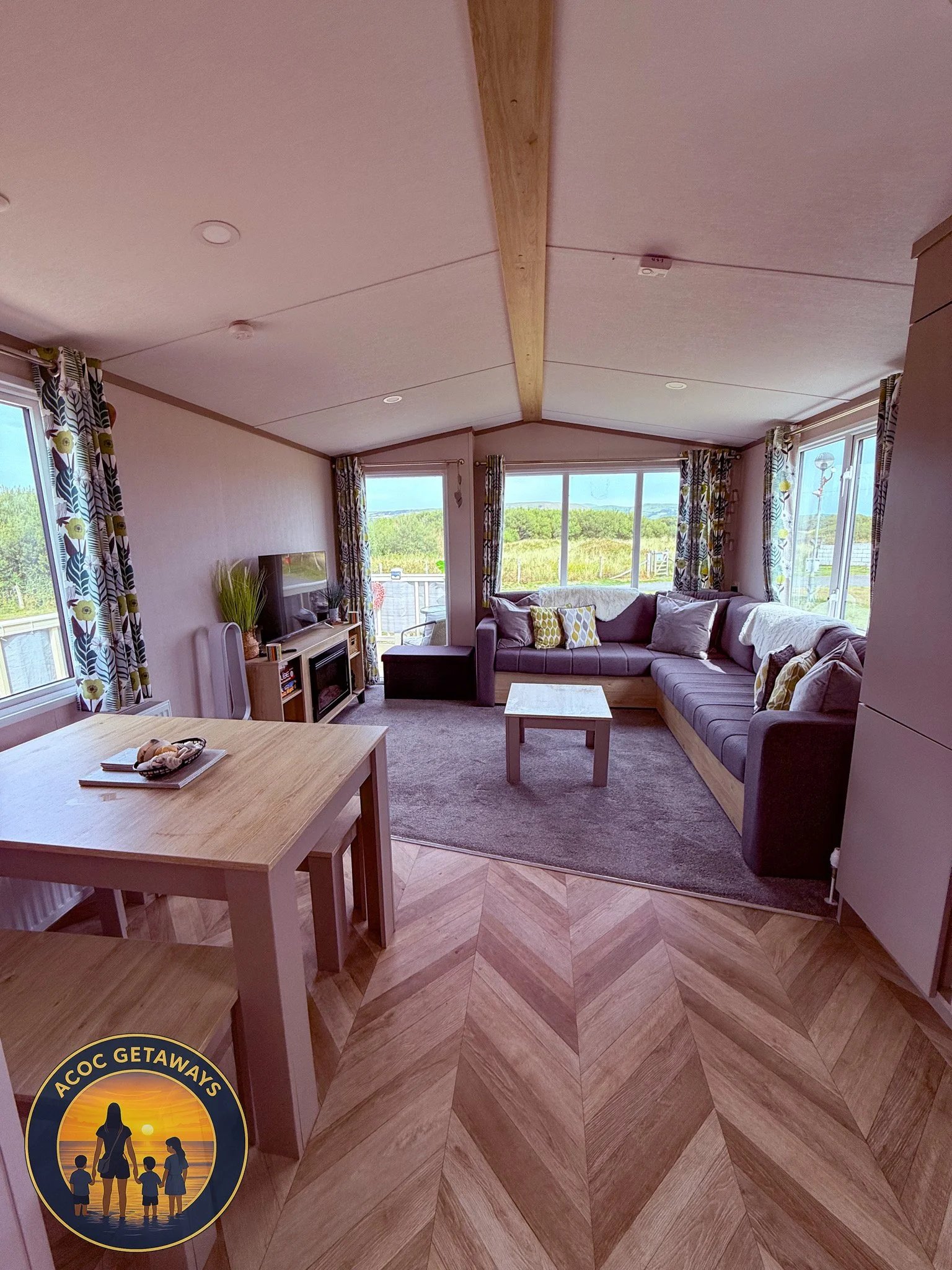 Living room with large windows, L-shaped sofa, coffee table, TV, curtains, and a view of greenery outside.
