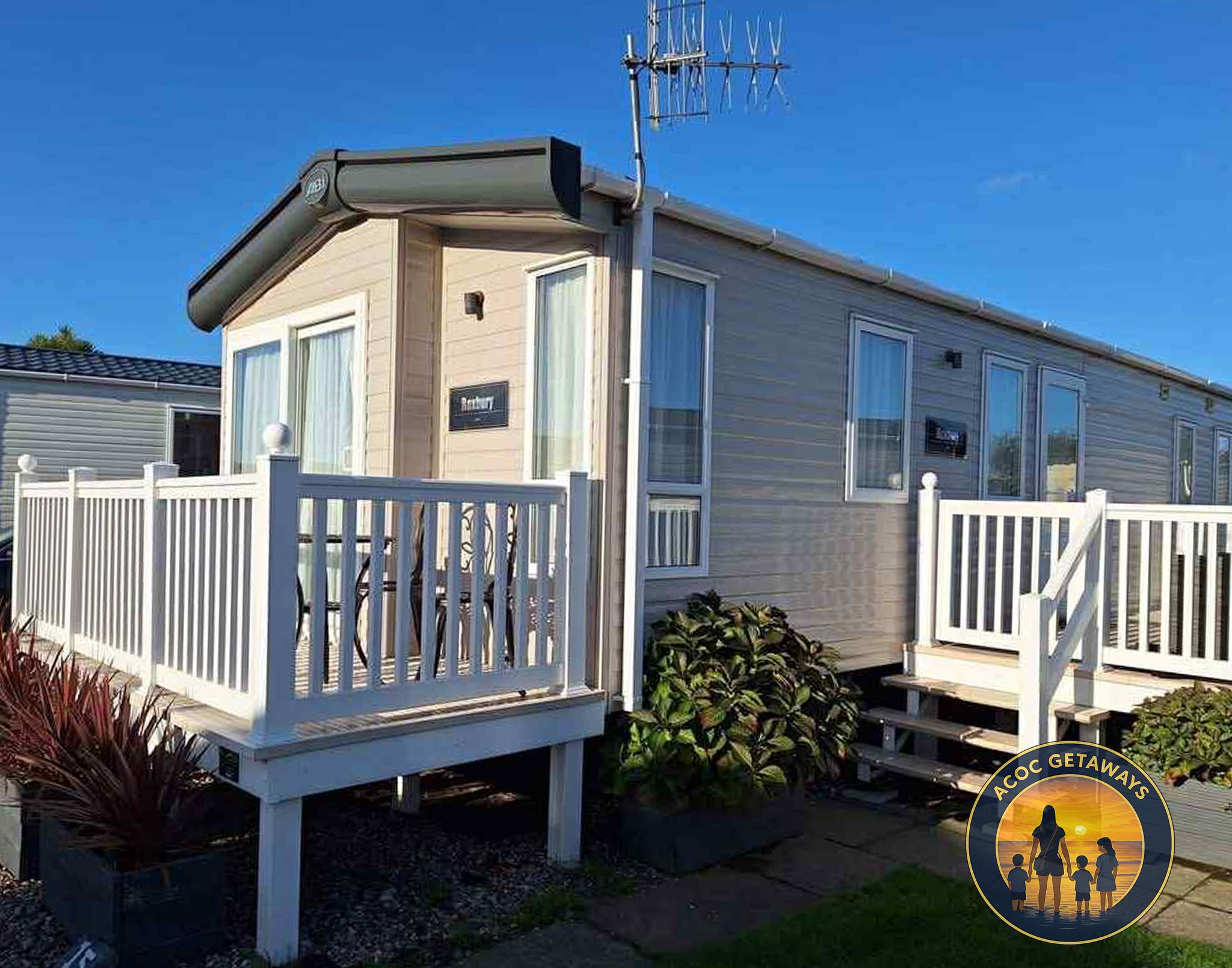 A beige mobile home with white trim and four windows, a small deck with white railing and steps, plants in a garden bed, and a TV antenna on the roof, under a clear blue sky.