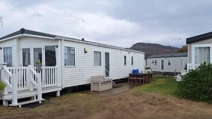 A row of white mobile homes with small porches, set on grassy and muddy ground, with cloudy sky and hills in the background.