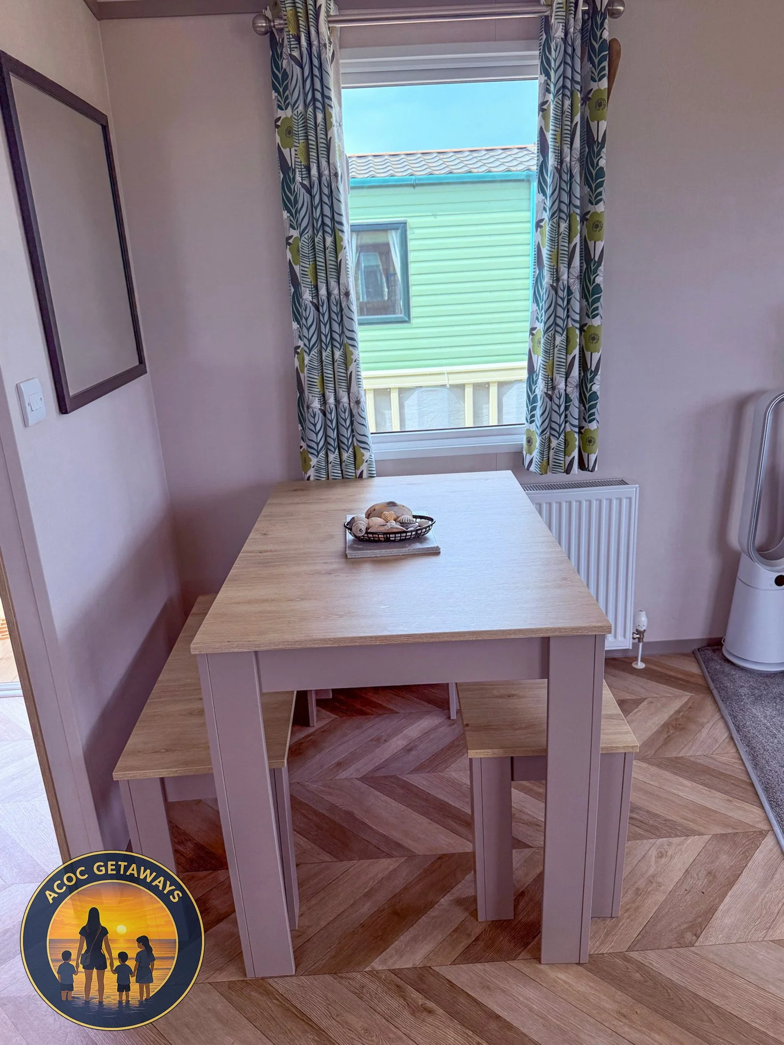 A wooden dining table with two matching benches placed next to a window with patterned curtains. Outside the window, there is a green building and clear blue sky. On the table, a small basket with shells is visible, and a white fan or air purifier is