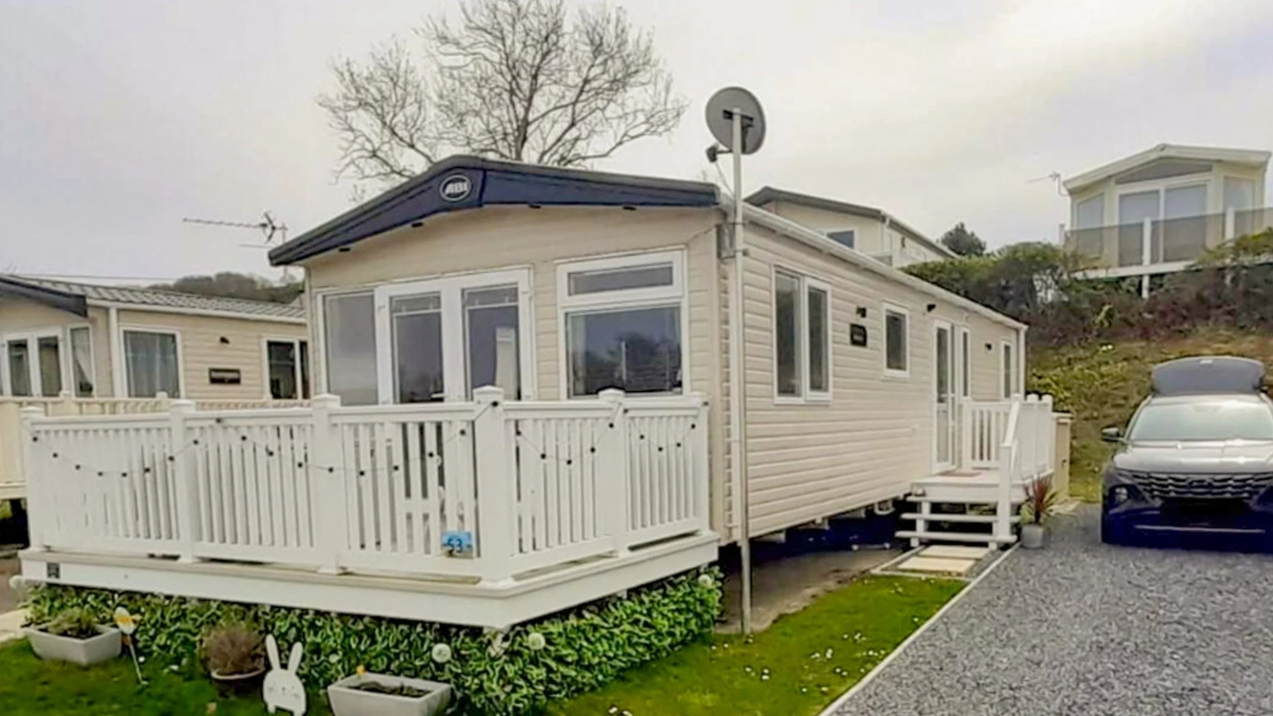 A beige mobile home with white trim and a small porch, surrounded by a white railing and decorative plants, with a blue car parked on a gravel driveway to the right.